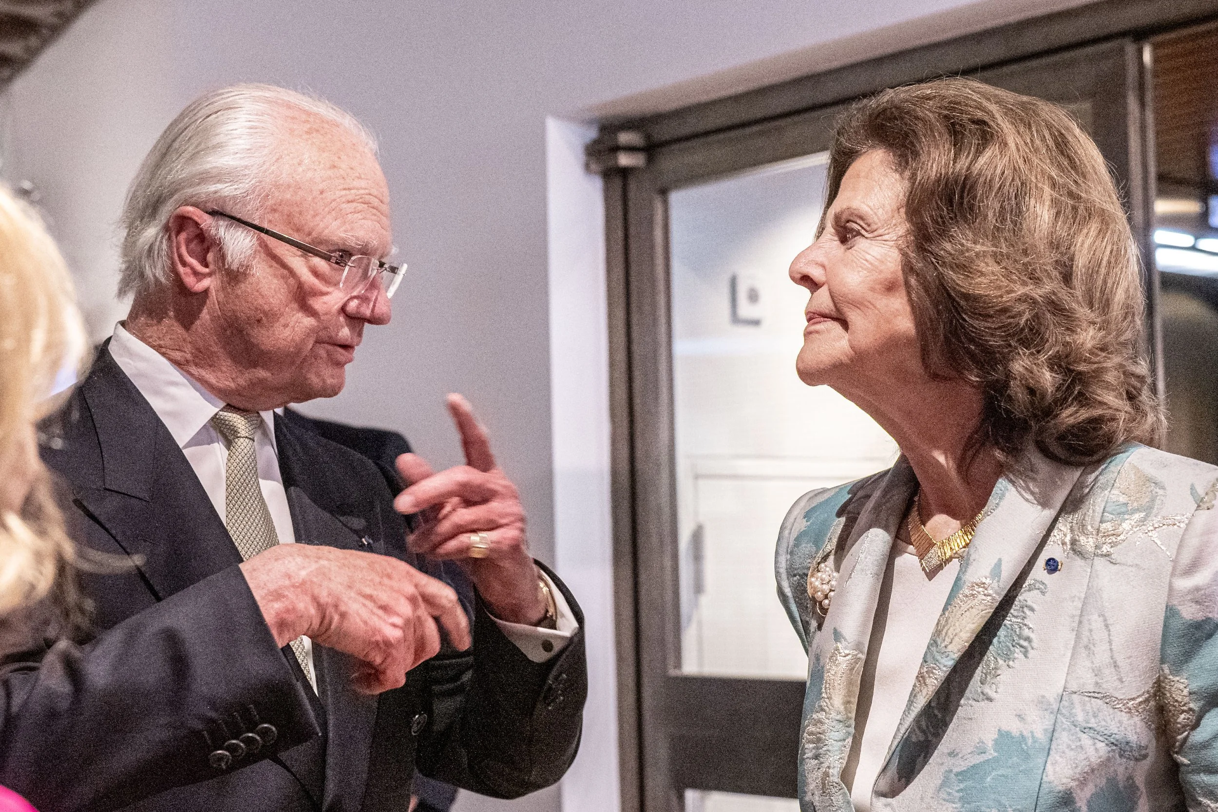 An elderly man with glasses and a woman with short hair and a light-colored patterned jacket engaged in conversation indoors.