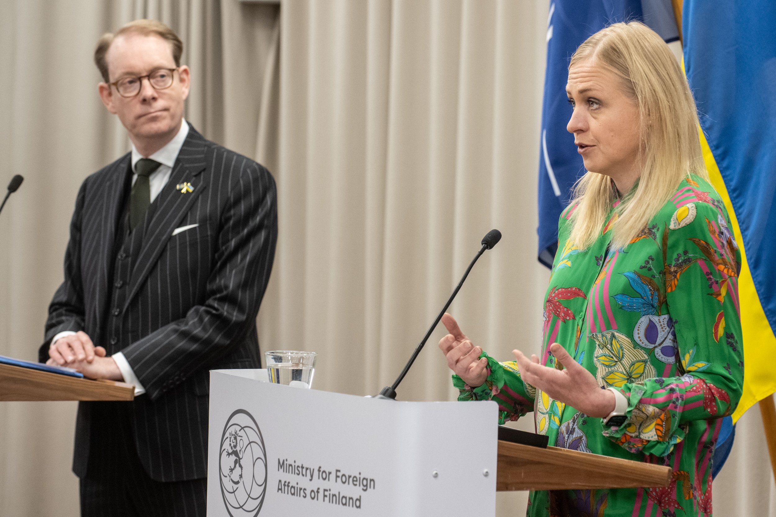 A woman speaking at a podium with a microphone, wearing a colorful, patterned dress, with flags and a man in a pinstripe suit standing nearby, during an event at the Ministry for Foreign Affairs of Finland.