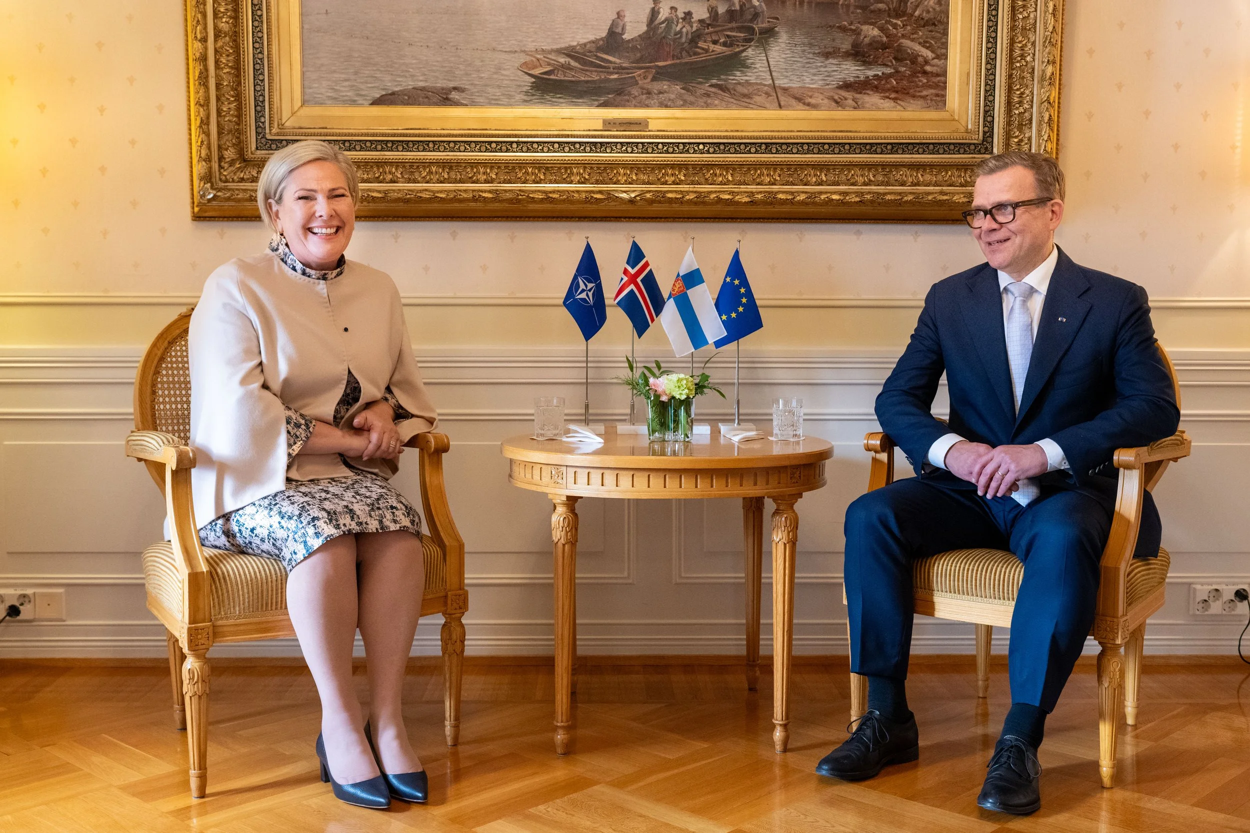 Two people sitting in chairs across from each other in a formal meeting room, with flags of NATO, Iceland, Finland, and the European Union on a small table between them, with a large framed painting above.