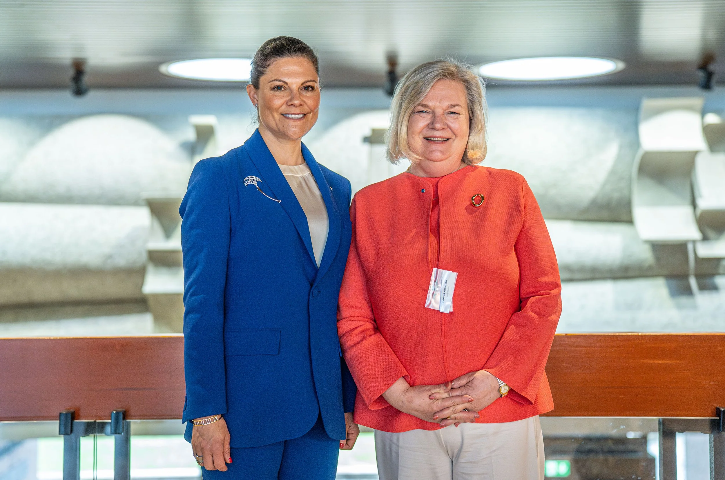 Two women standing together, smiling, in a modern indoor setting. One woman wears a blue suit with a pin, and the other wears an orange jacket with a brooch. They are positioned in front of a wooden railing, with an abstract architectural background.
