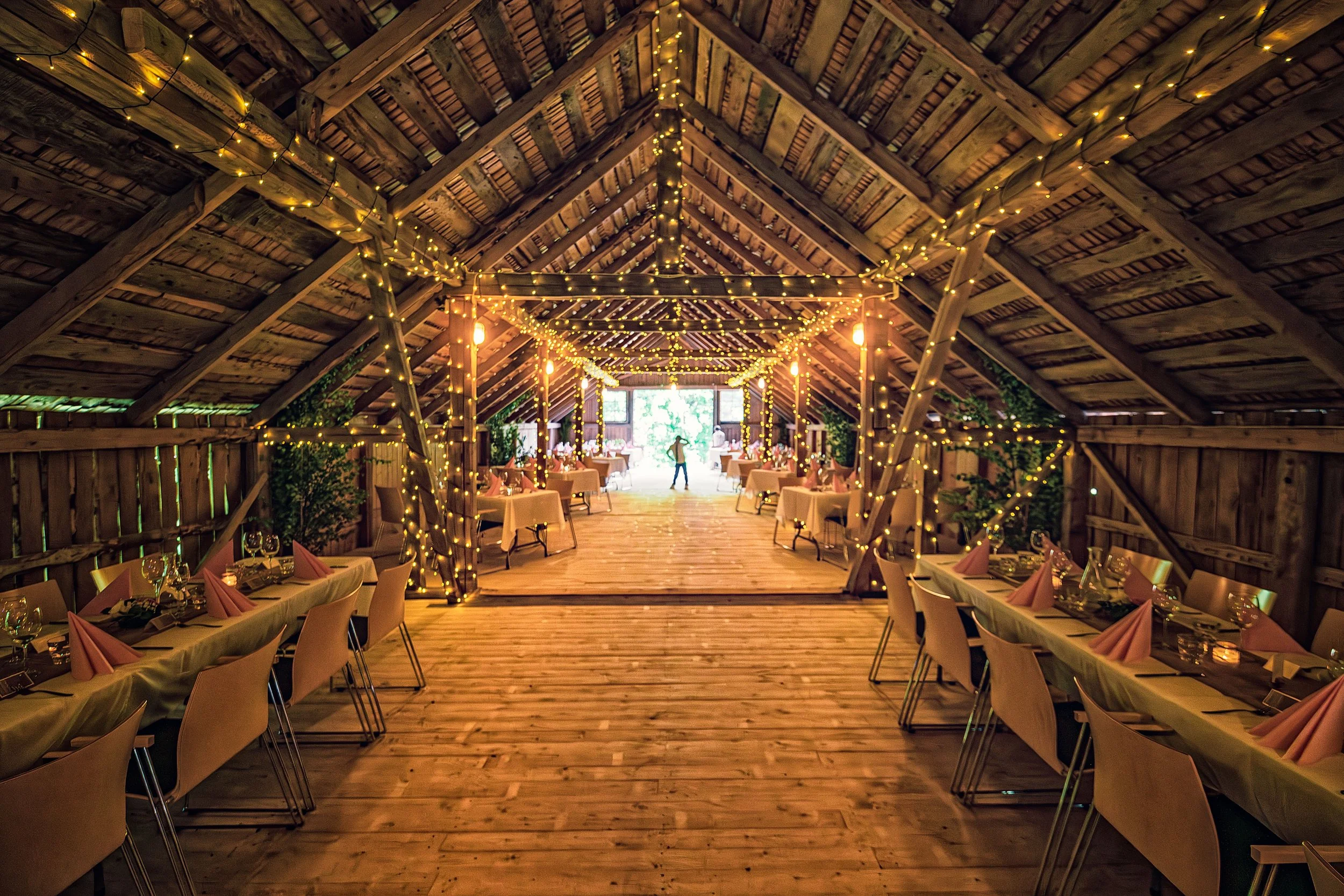Interior of a wooden barn decorated with string lights hanging from the ceiling and wrapped around support beams. Long dining tables with pink napkins, glassware, and cutlery are set along both sides of the open space, leading to a bright doorway at the far end with a person standing in front of it.