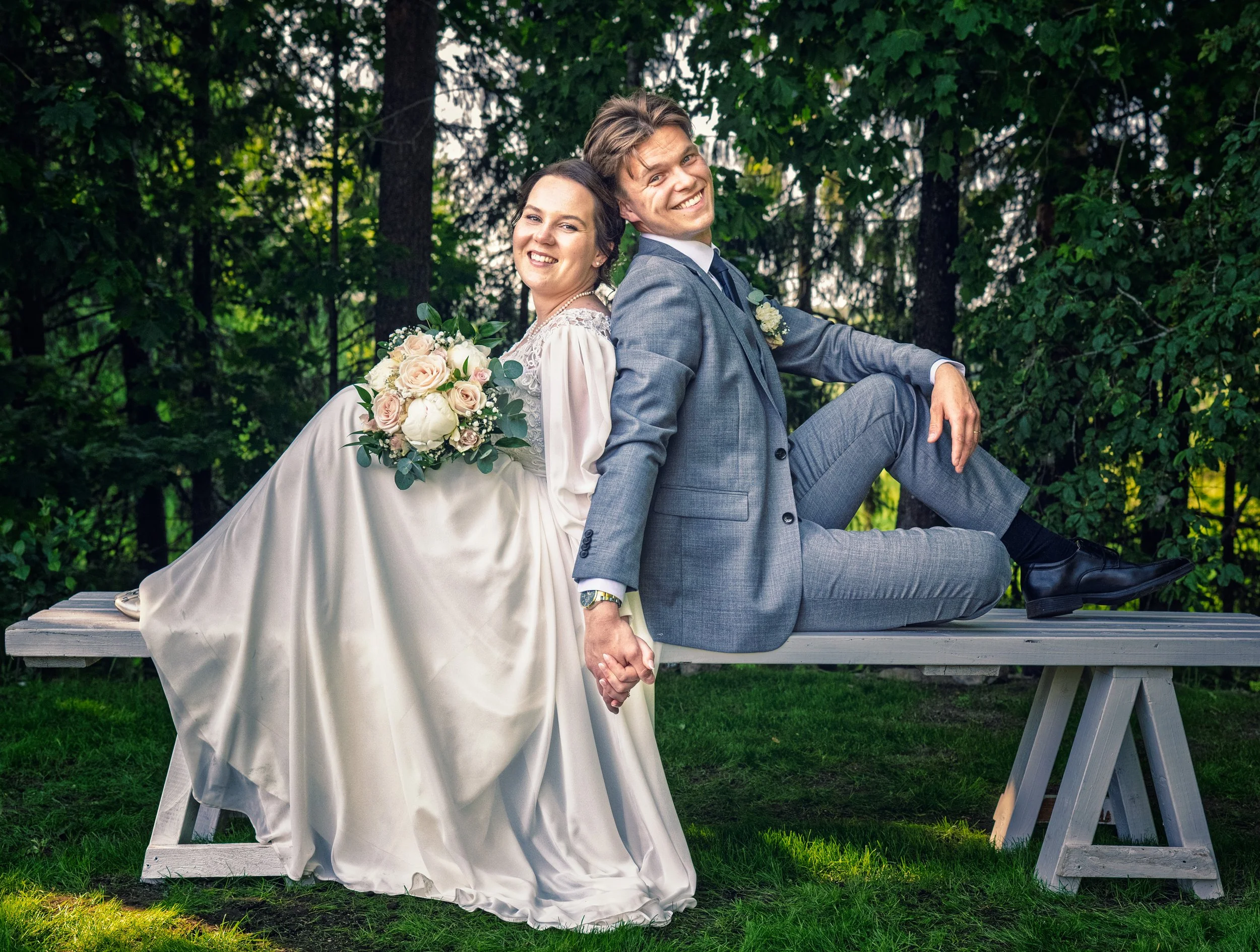 A bride and groom sitting back-to-back on a wooden bench outdoors, holding hands. The bride is wearing a white wedding dress and holding a bouquet of flowers. The groom is wearing a gray suit. Both are smiling. The background features green trees and foliage.