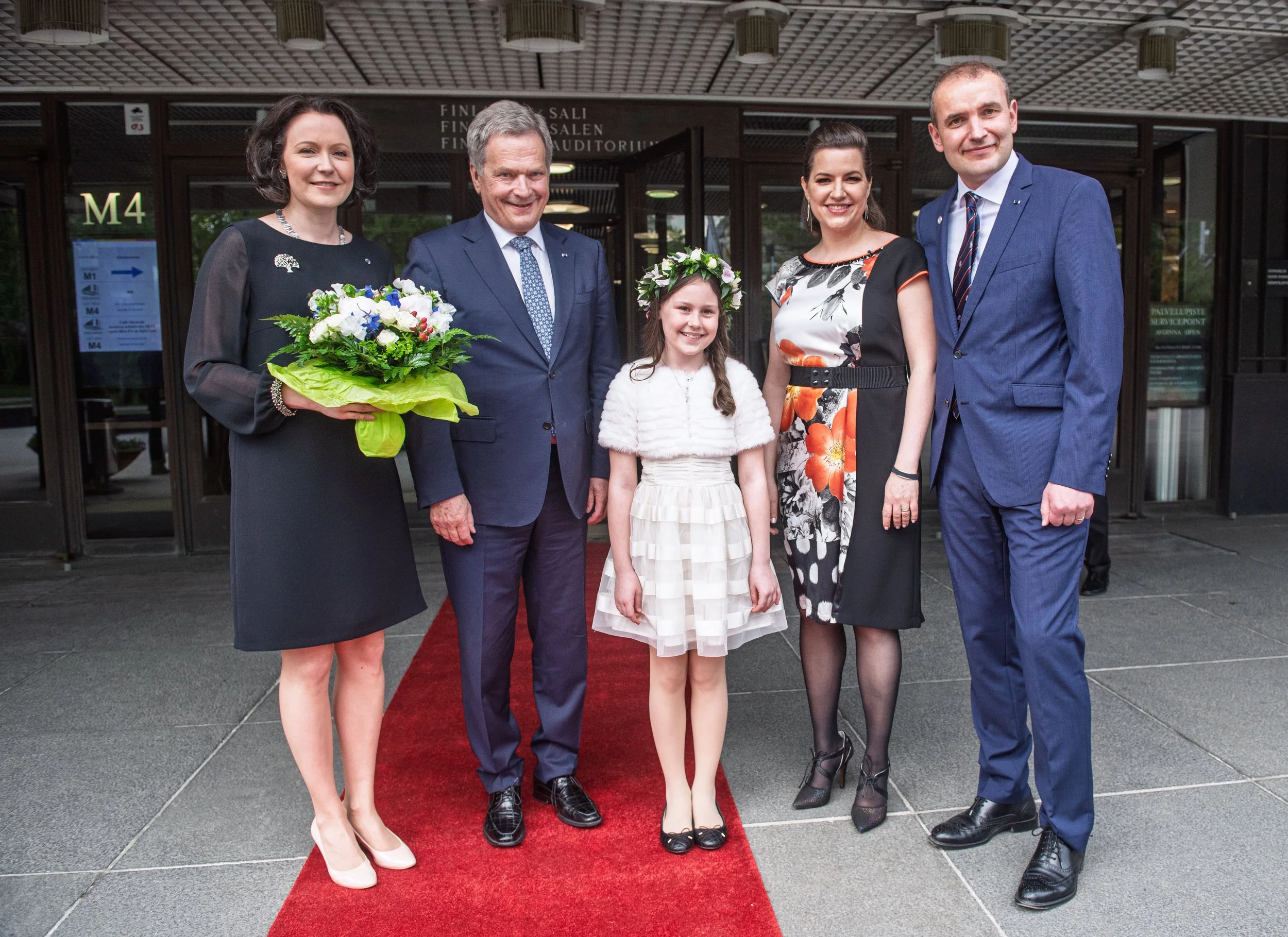 Group of five people standing on a red carpet in front of a building, dressed in formal attire, with a woman holding a bouquet of flowers and a young girl wearing a floral headband in the center.