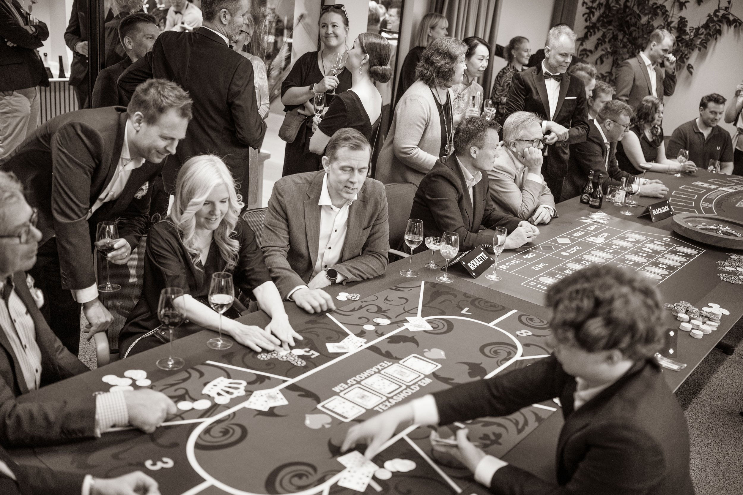 People sitting and standing around a casino table playing a game, with drinks in hand, some watching and others actively participating, in a social gathering or party setting.