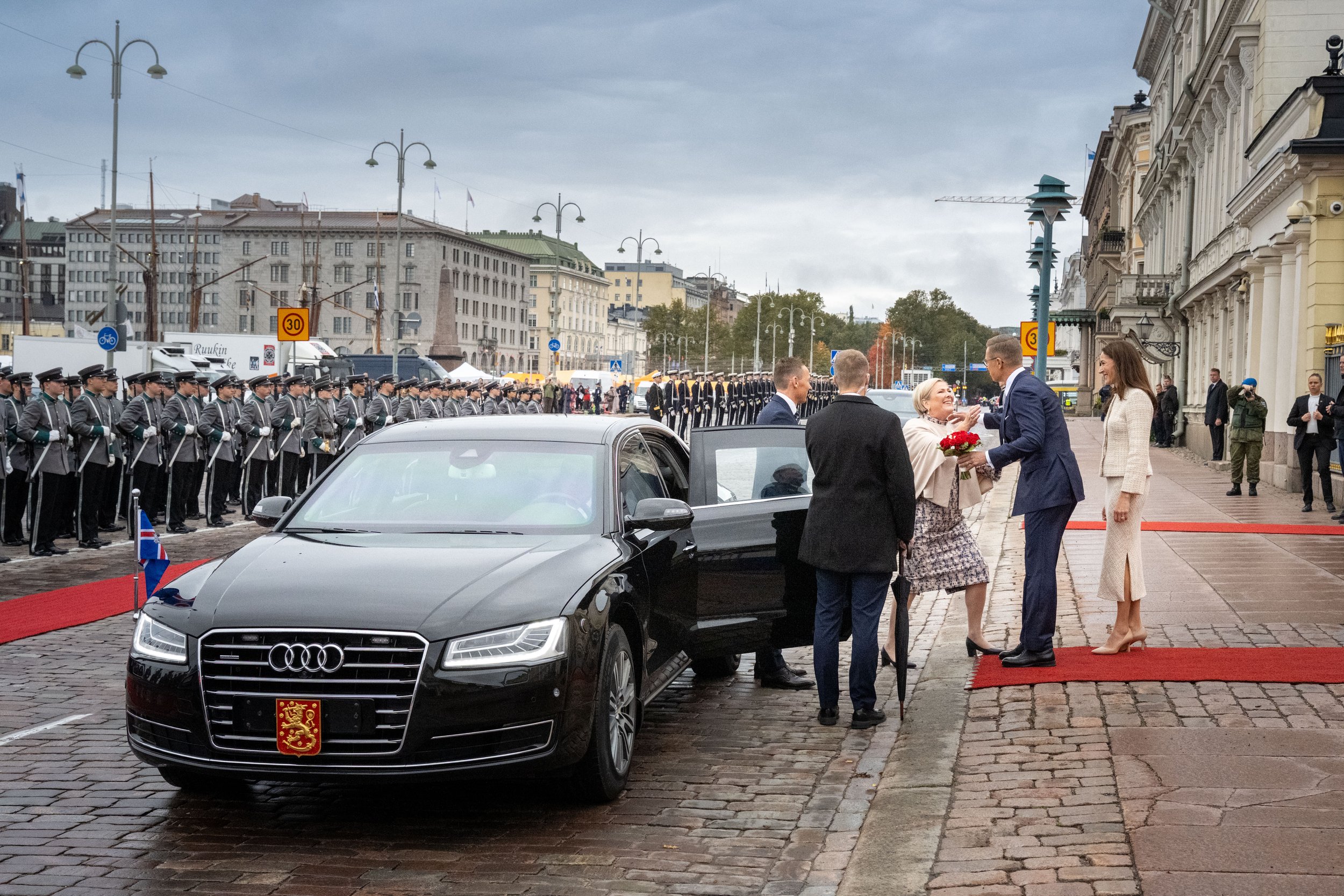 A formal outdoor ceremony with a black Audi car on a cobblestone street, surrounded by well-dressed people, including a woman receiving flowers, along with soldiers standing in formation in the background.