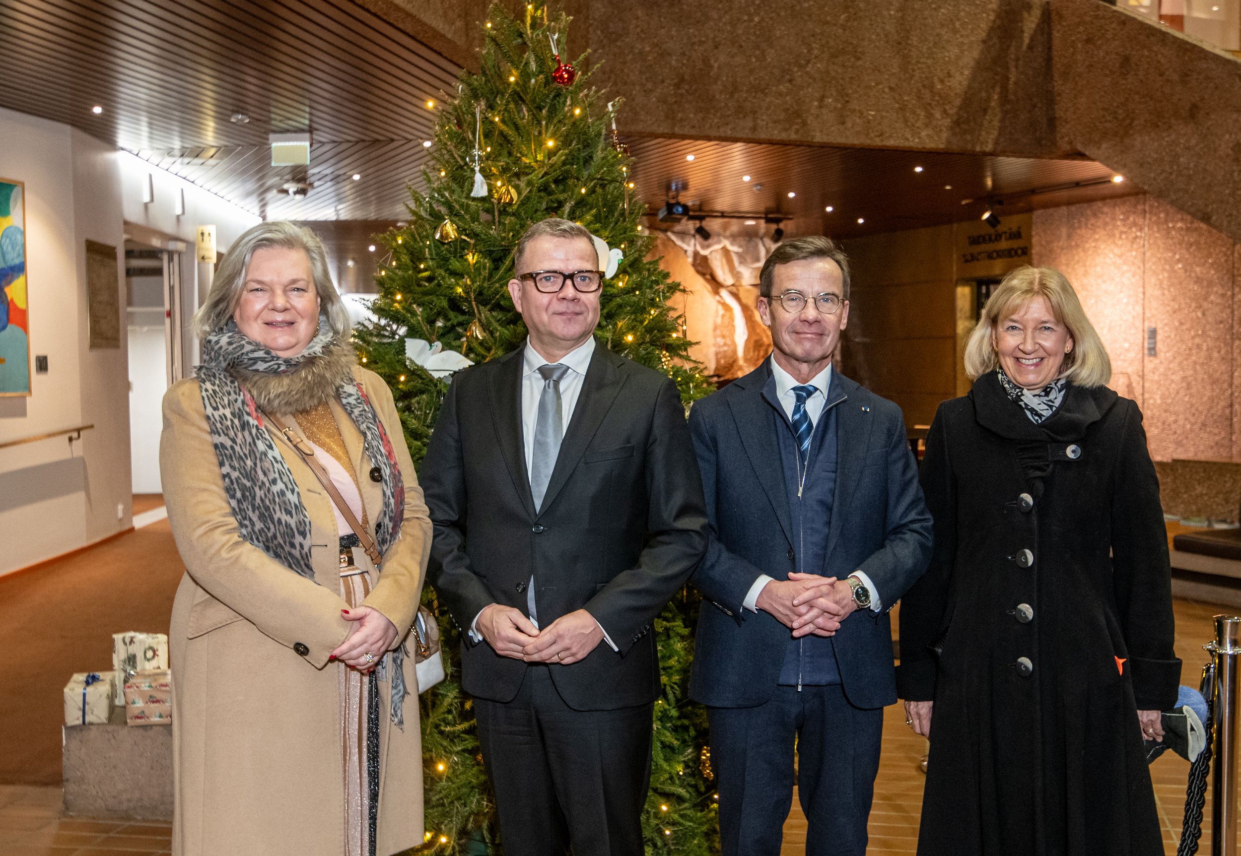 Four people standing in front of a decorated Christmas tree indoors, dressed in winter clothing, with gift boxes on the floor to the left.