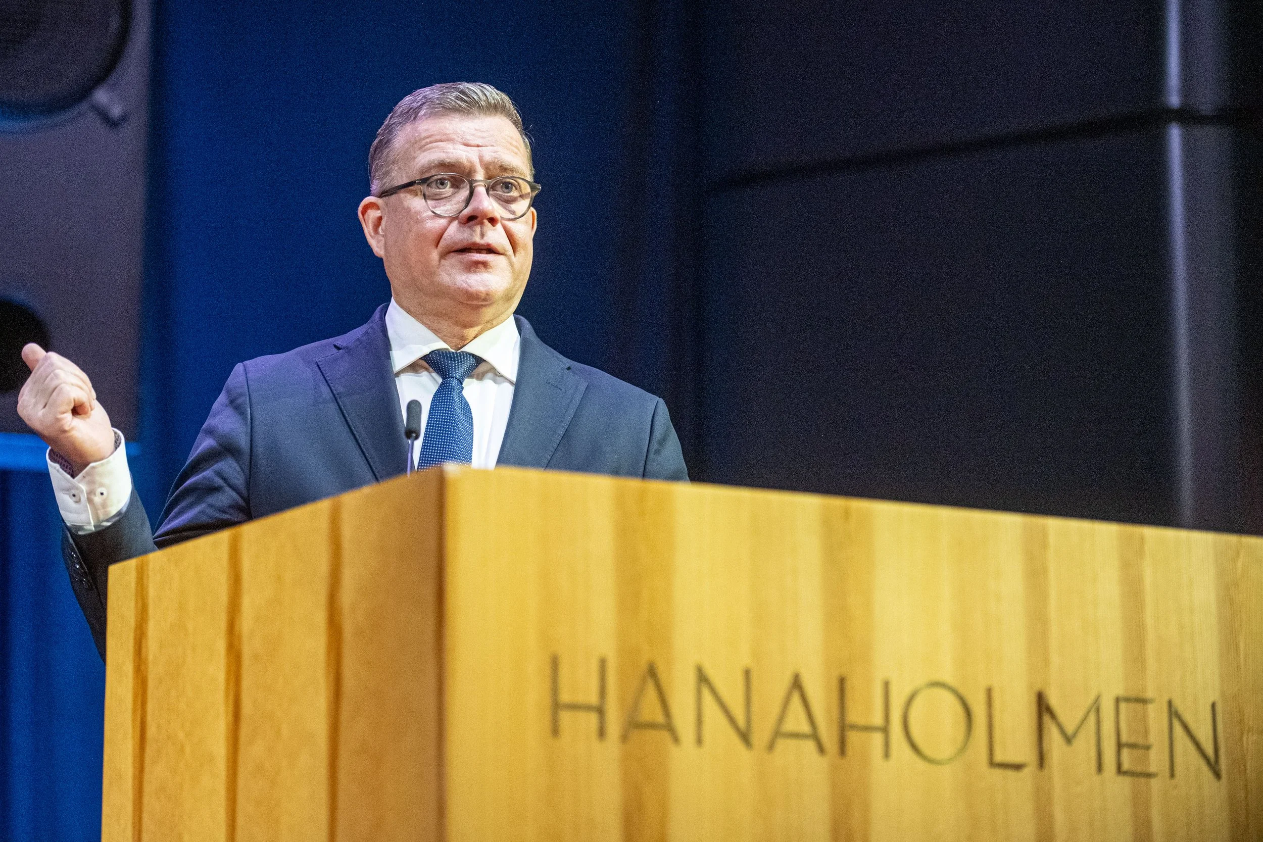 A man in a suit and glasses speaking at a podium with the words 'HANAHOLMEN' written on it, in a formal setting.