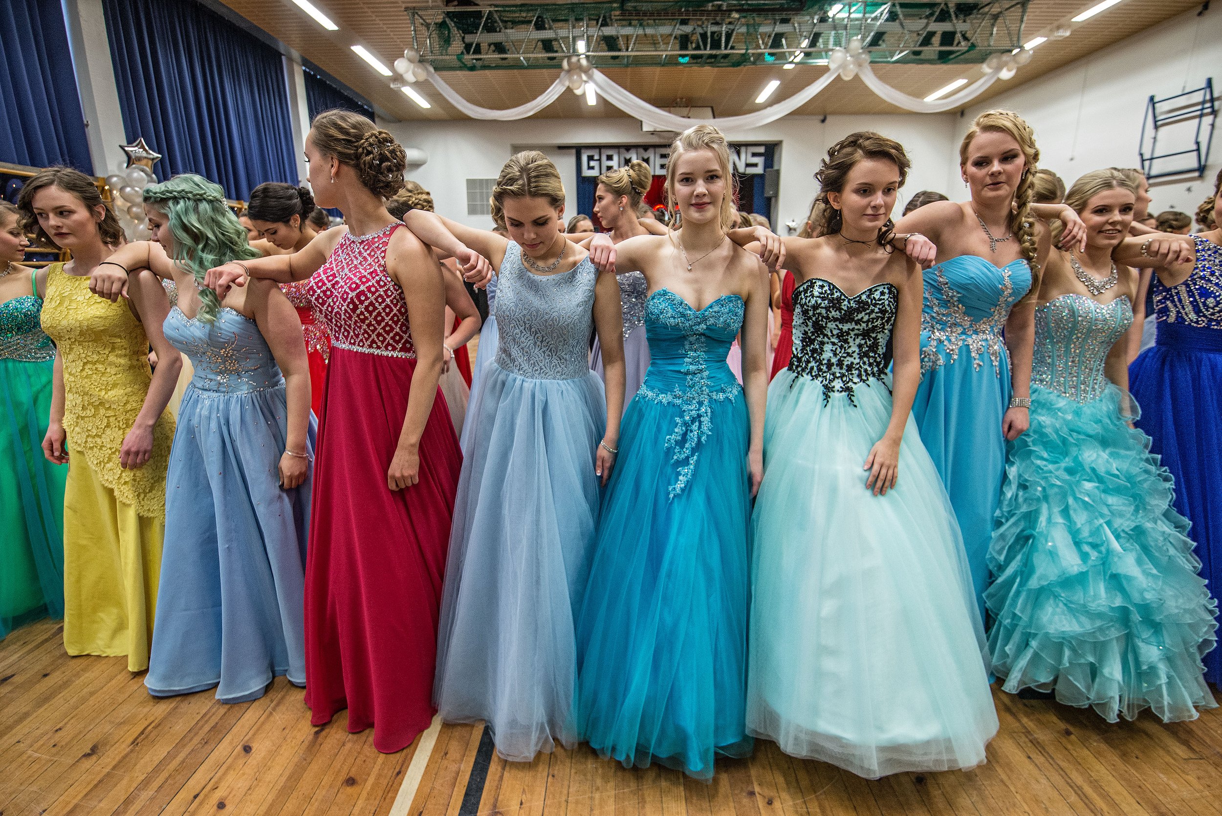 Group of young women in colorful evening gowns standing in a gymnasium, arms linked together in a line, participating in a formal event or dance.