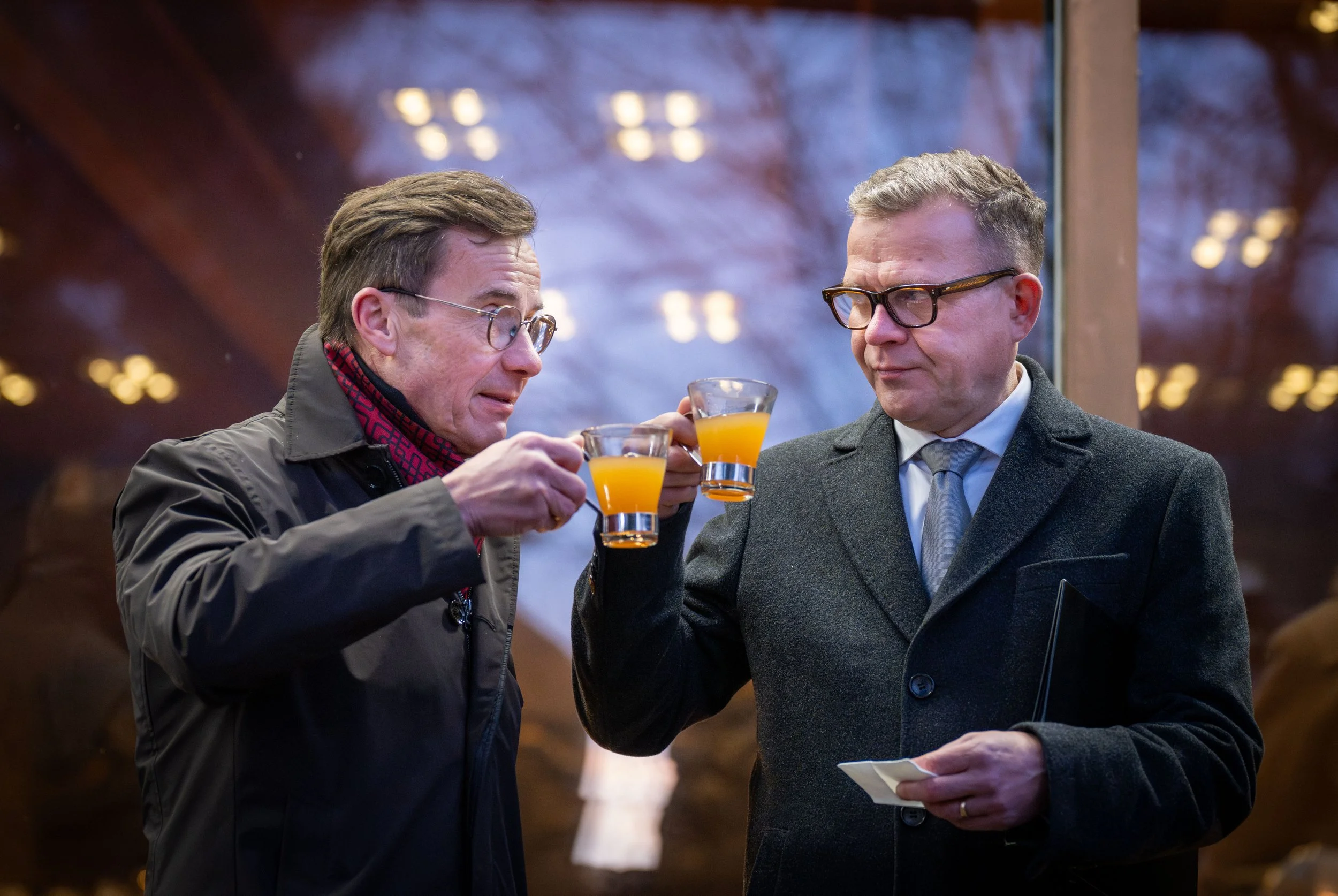 Two men wearing glasses and suits, holding glasses of orange juice in a toast, standing indoors near a window with a blurred outdoor scene.