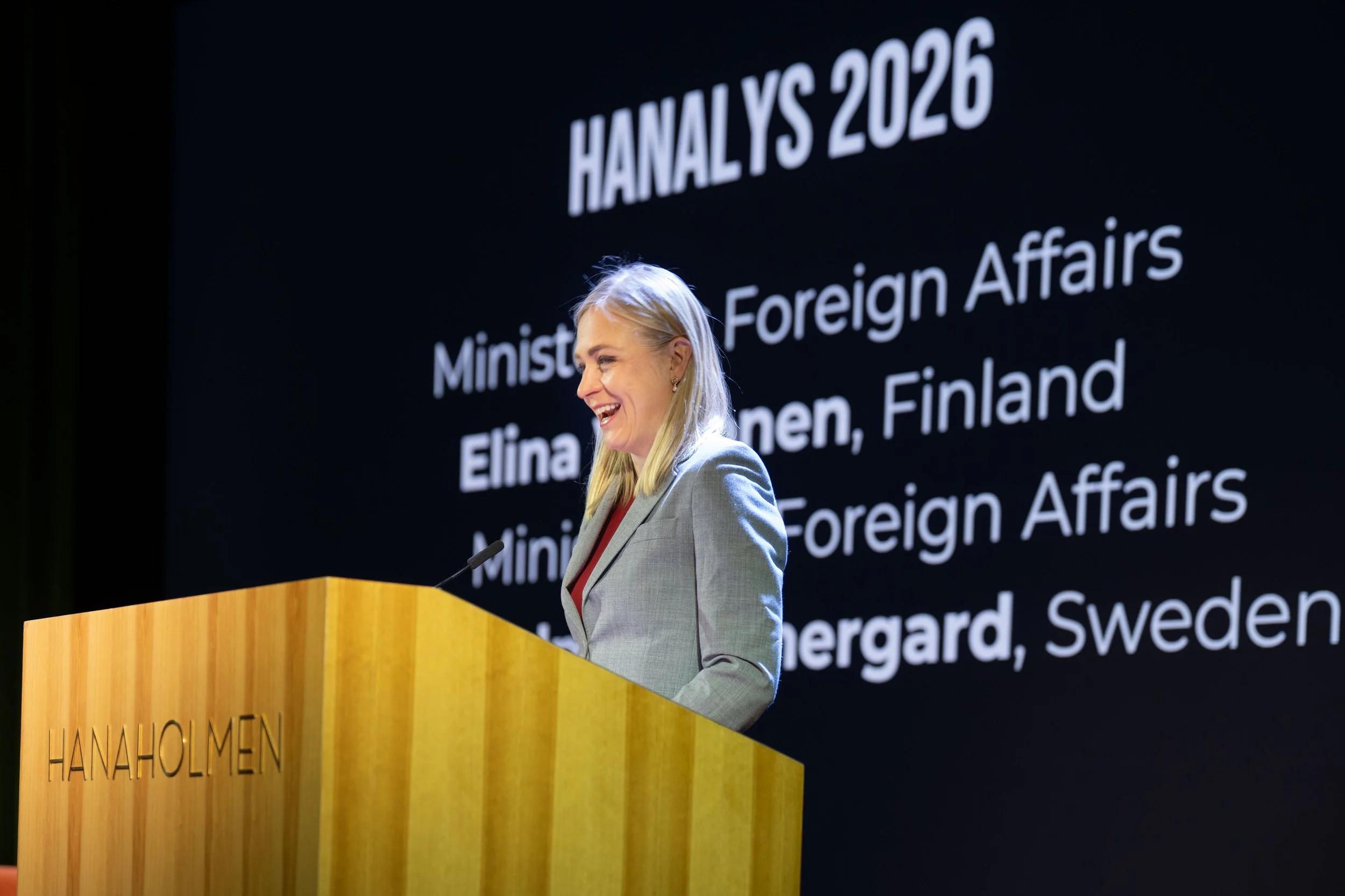 A woman stands at a wooden podium labeled 'HANAHOLMEN,' smiling and speaking into a microphone. Behind her is a black screen displaying the text 'HANALYS 2026,' along with information about the Finnish Ministry of Foreign Affairs, Elina Korpinen from