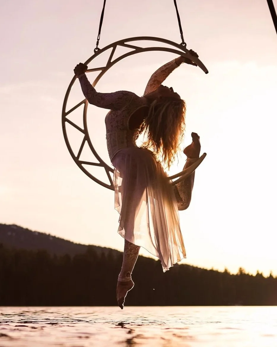A woman performing an aerial dance or acrobatics on a crescent moon-shaped aerial hoop, silhouetted against a sunset or sunrise over a body of water and distant hills.