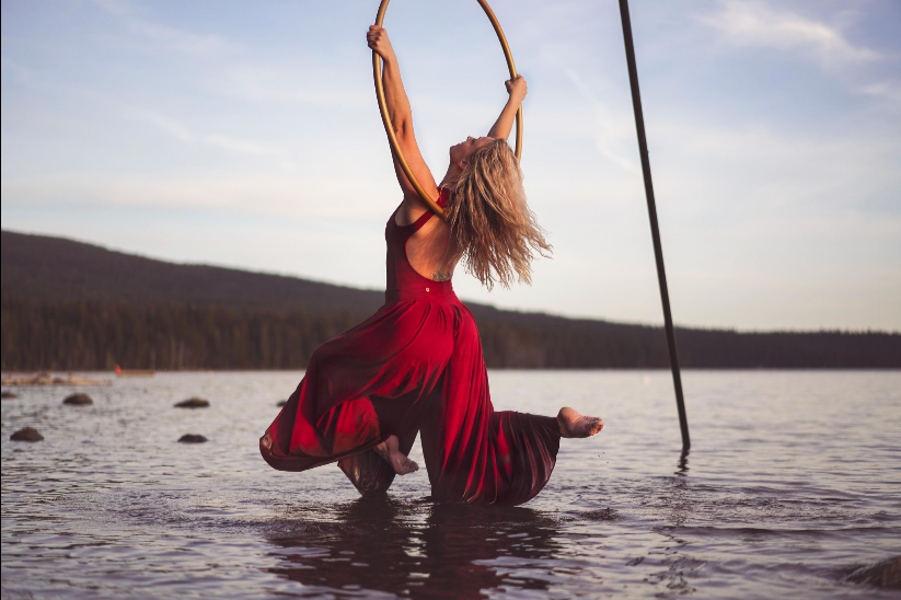 A woman in a red dress kneels in water, holding a hoop above her head, with a scenic lake and distant forest in the background.