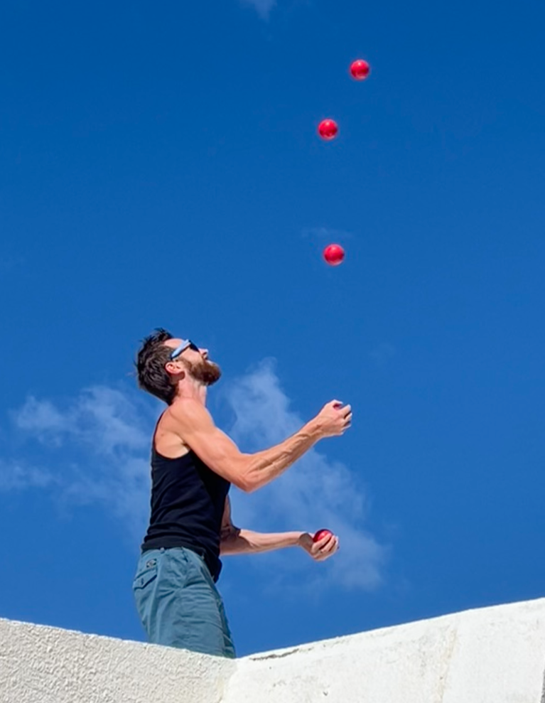 A man with sunglasses, a beard, and a sleeveless black shirt is juggling three red balls outdoors against a bright blue sky.