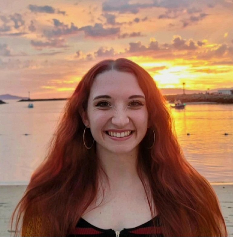 A young woman with long red hair, wearing hoop earrings and a black top, smiling at sunset by the water with boats in the background.
