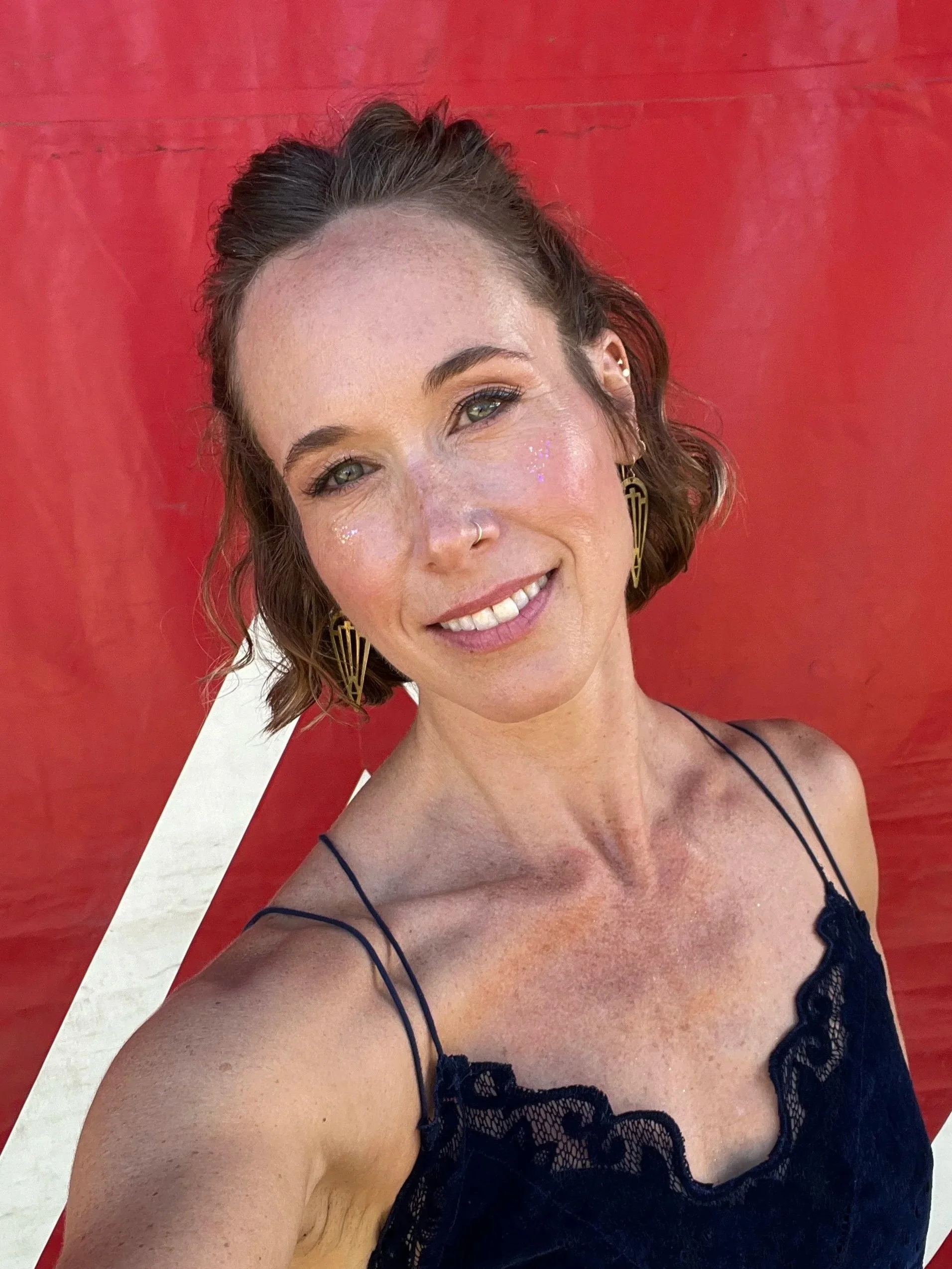 A woman with short wavy brown hair, light skin, and green eyes smiling at the camera, wearing gold earrings and a black lace spaghetti strap top, standing in front of a red backdrop.
