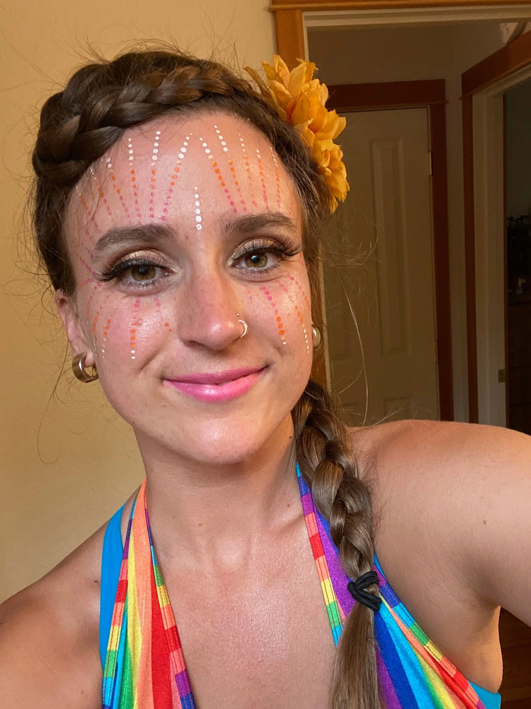 A woman with makeup and face paint, wearing rainbow-colored straps, with a yellow flower in her braided hair, smiling indoors.