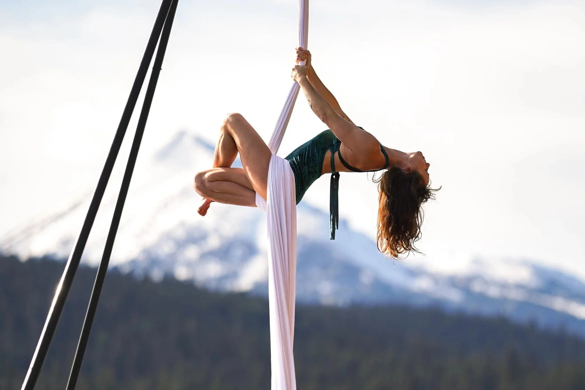 A woman performing an aerial silk routine outdoors with snow-capped mountains in the background.