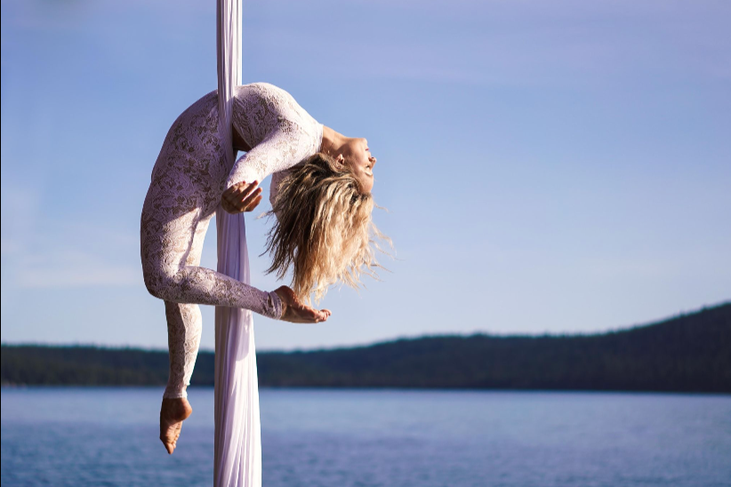A woman performing aerial silk yoga outdoors near a body of water with hills in the background.