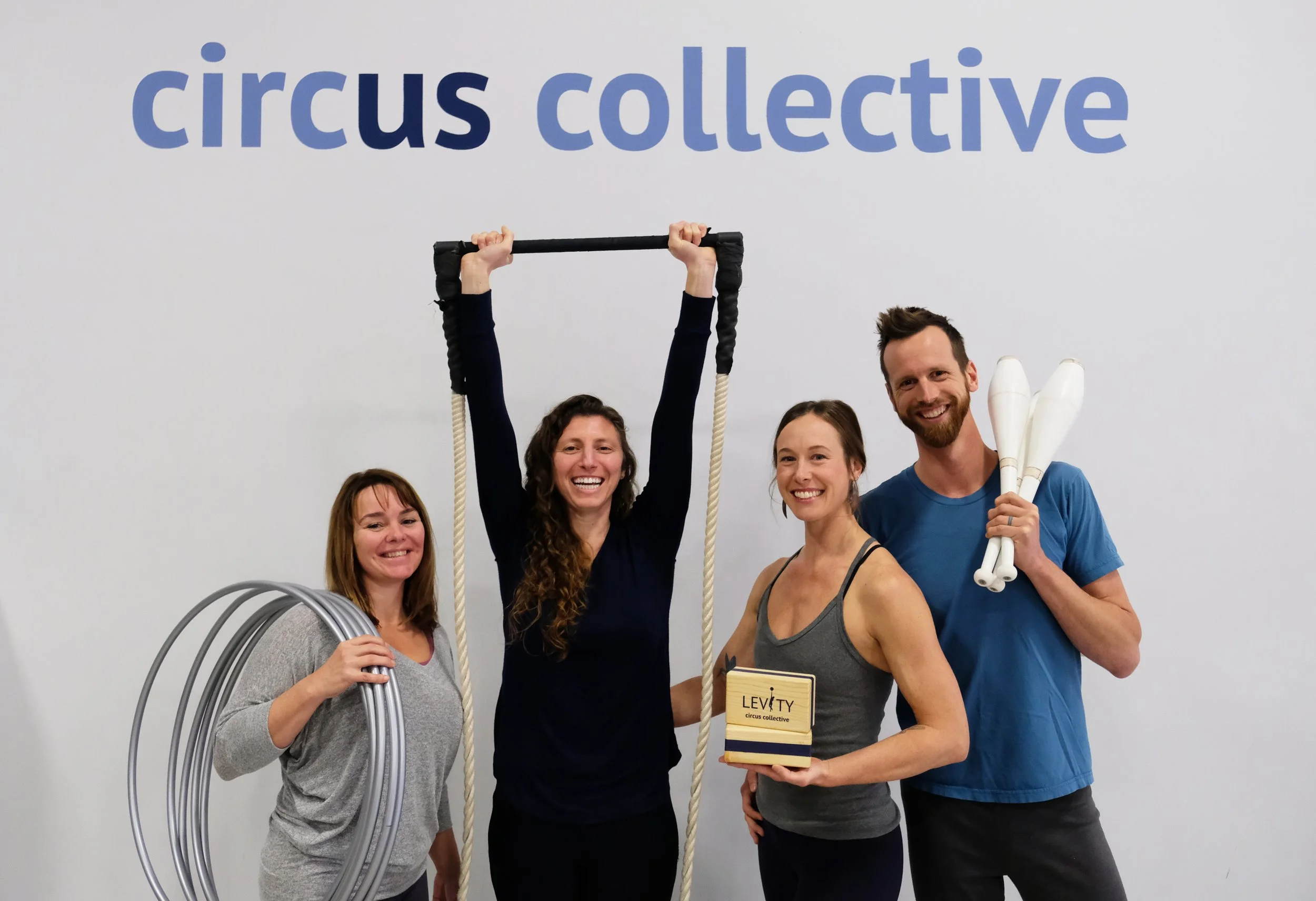 Group of four happy circus performers holding various circus props in front of a white wall with the words 'circus collective' in blue letters.