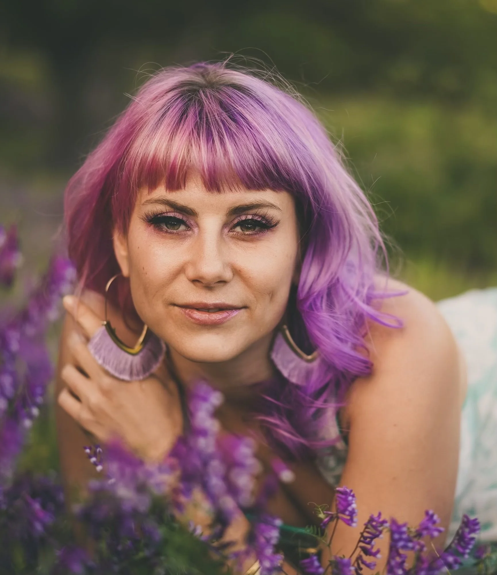 A woman with purple and pink hair lying in a field of purple flowers, facing the camera with a slight smile.