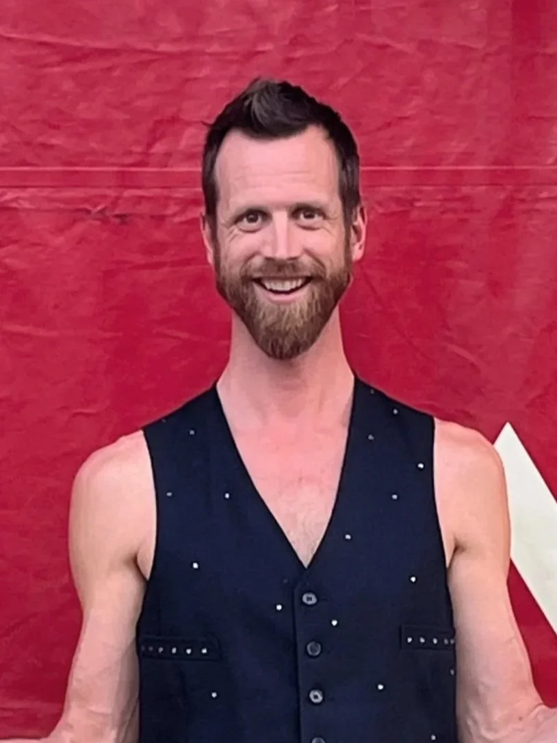 A smiling man with a beard and short brown hair, wearing a sleeveless black vest with small white dots, standing against a red background.