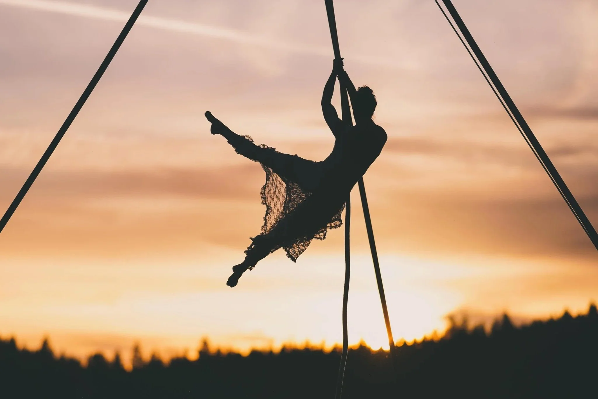 Silhouette of a woman performing an aerial acrobatic pose on silk at sunset with a blurred landscape in the background.