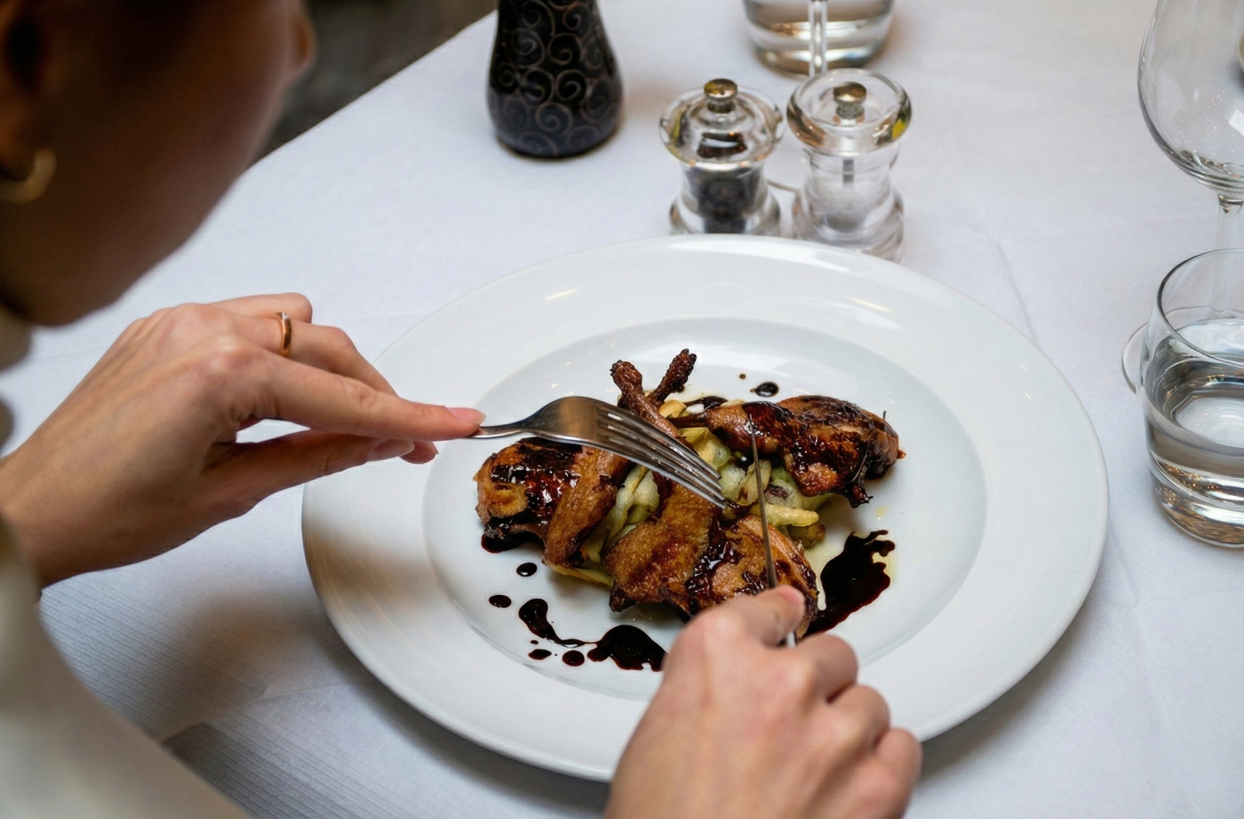 Person using a fork and knife to cut into a plate of roasted chicken with vegetables, drizzled with balsamic glaze, on a white table with water glasses and condiments.