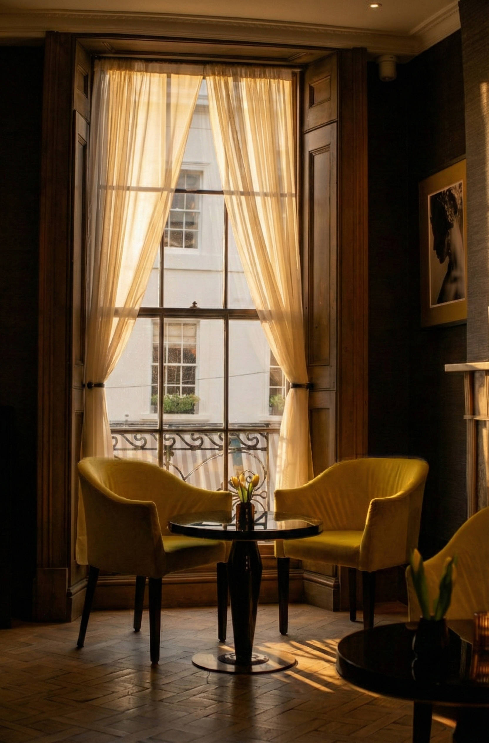 A cozy corner with two yellow upholstered chairs, a small round black table with a vase of yellow tulips, and a large window with cream-colored curtains letting in warm natural light.