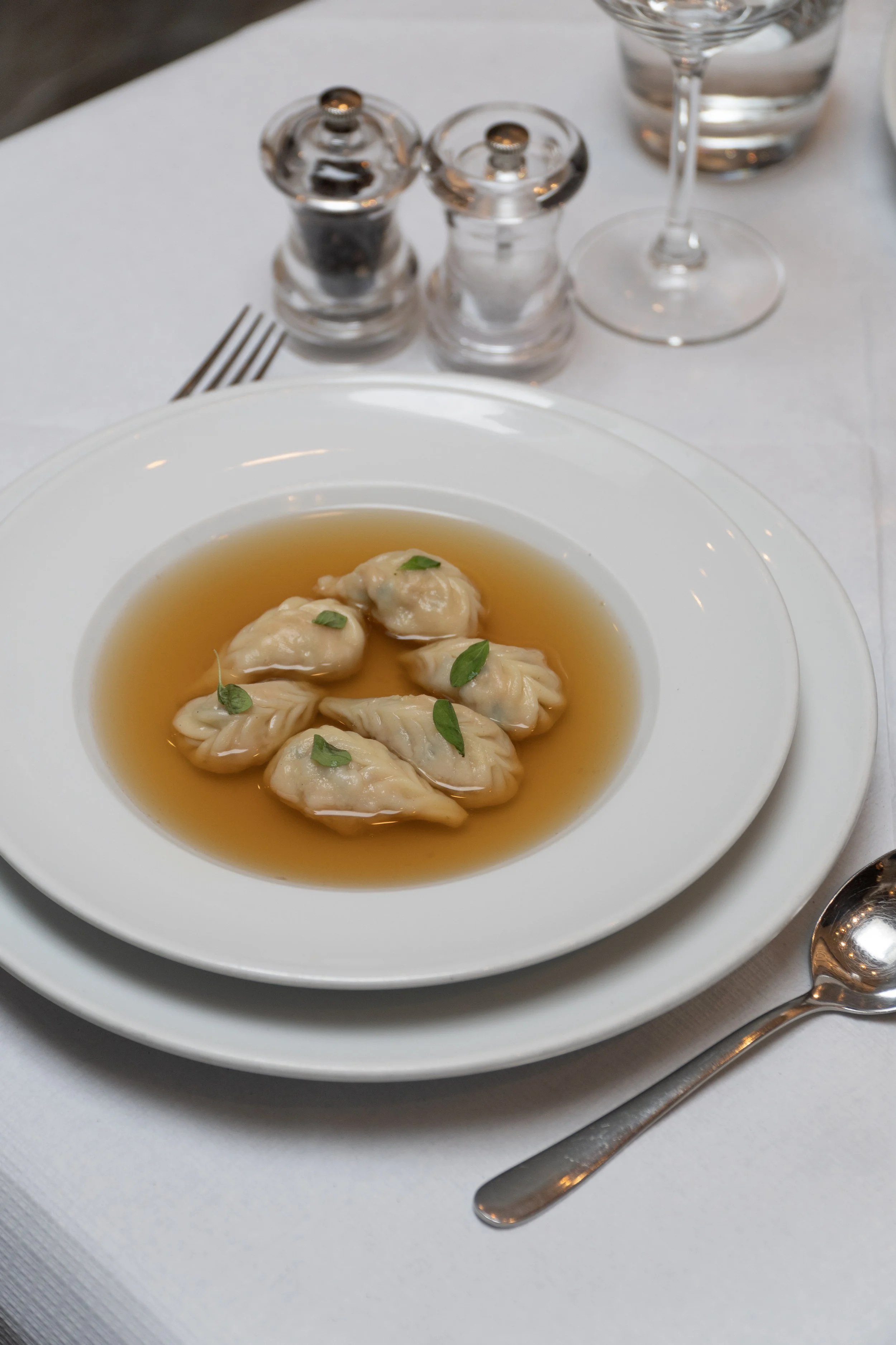 Plate of dumplings in broth garnished with small green leaves on a white tablecloth with salt and pepper shakers, a spoon, fork, and glass of water.