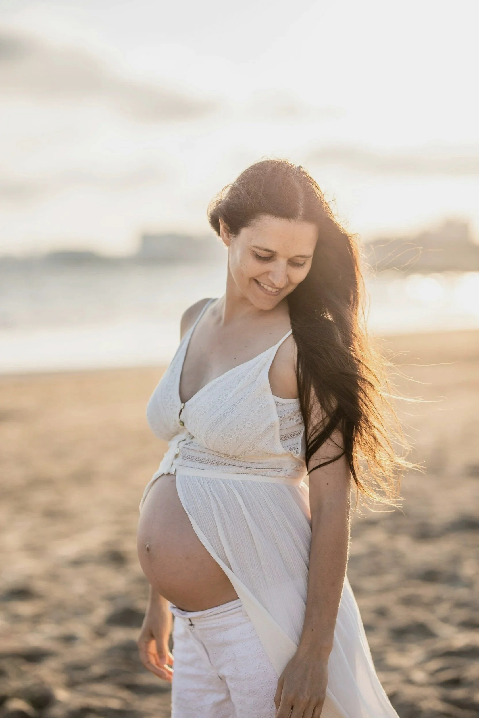 A pregnant woman with long dark hair, dressed in a white dress, standing on a beach during sunset, smiling and looking down. She is planning for her prenatal planning session with Julie from Integrative Lactation Services in Central Oregon.