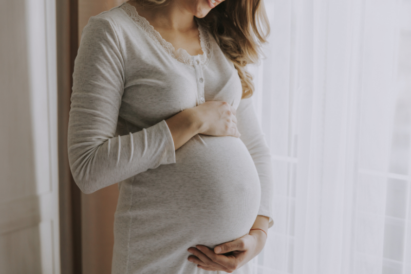 Pregnant woman gently touching her belly, standing near a window with sheer curtains in Bend Oregon.