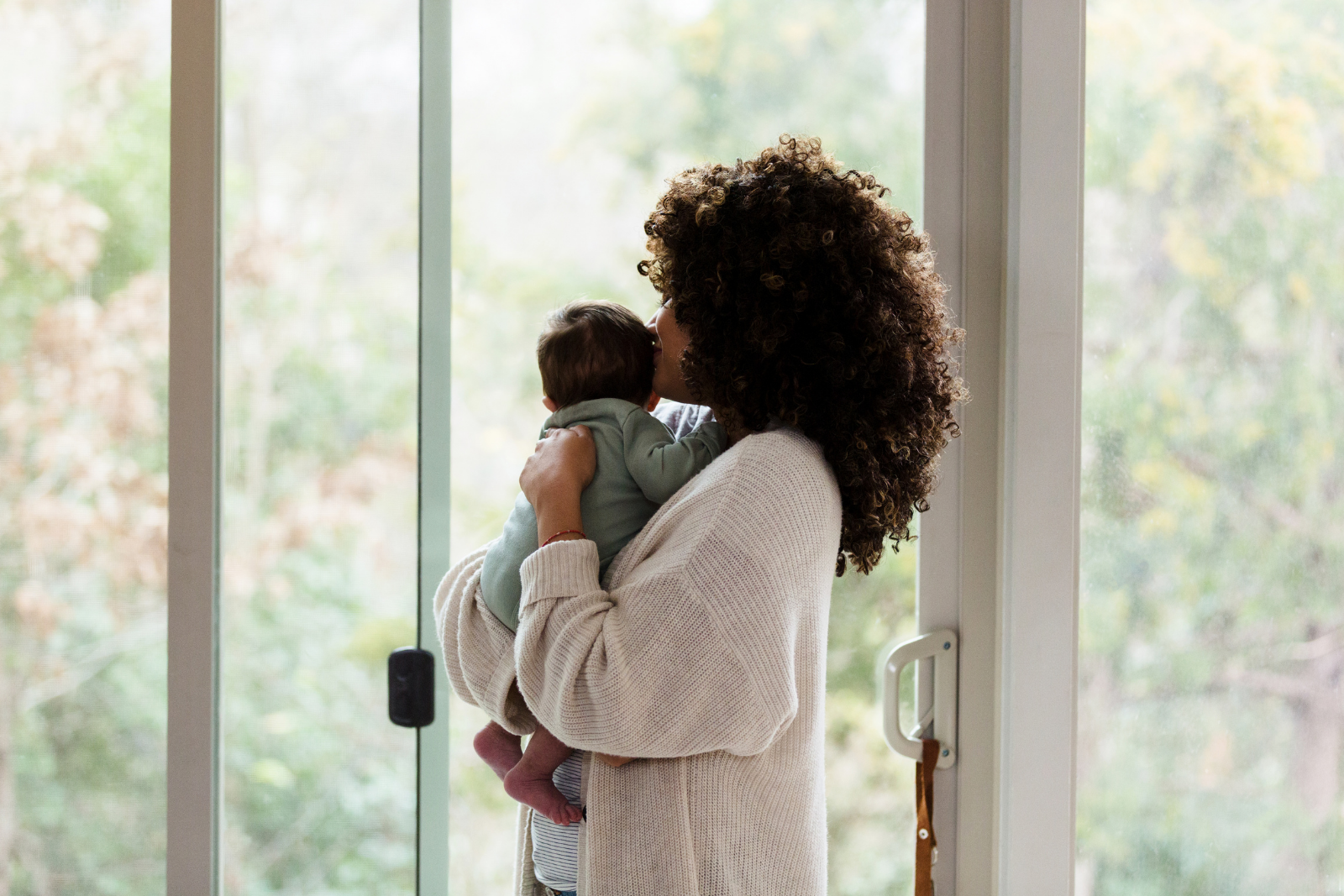 A woman with curly hair holding a baby near a sliding glass door after a calm, personalized breastfeeding support session in her Madras, Oregon, home