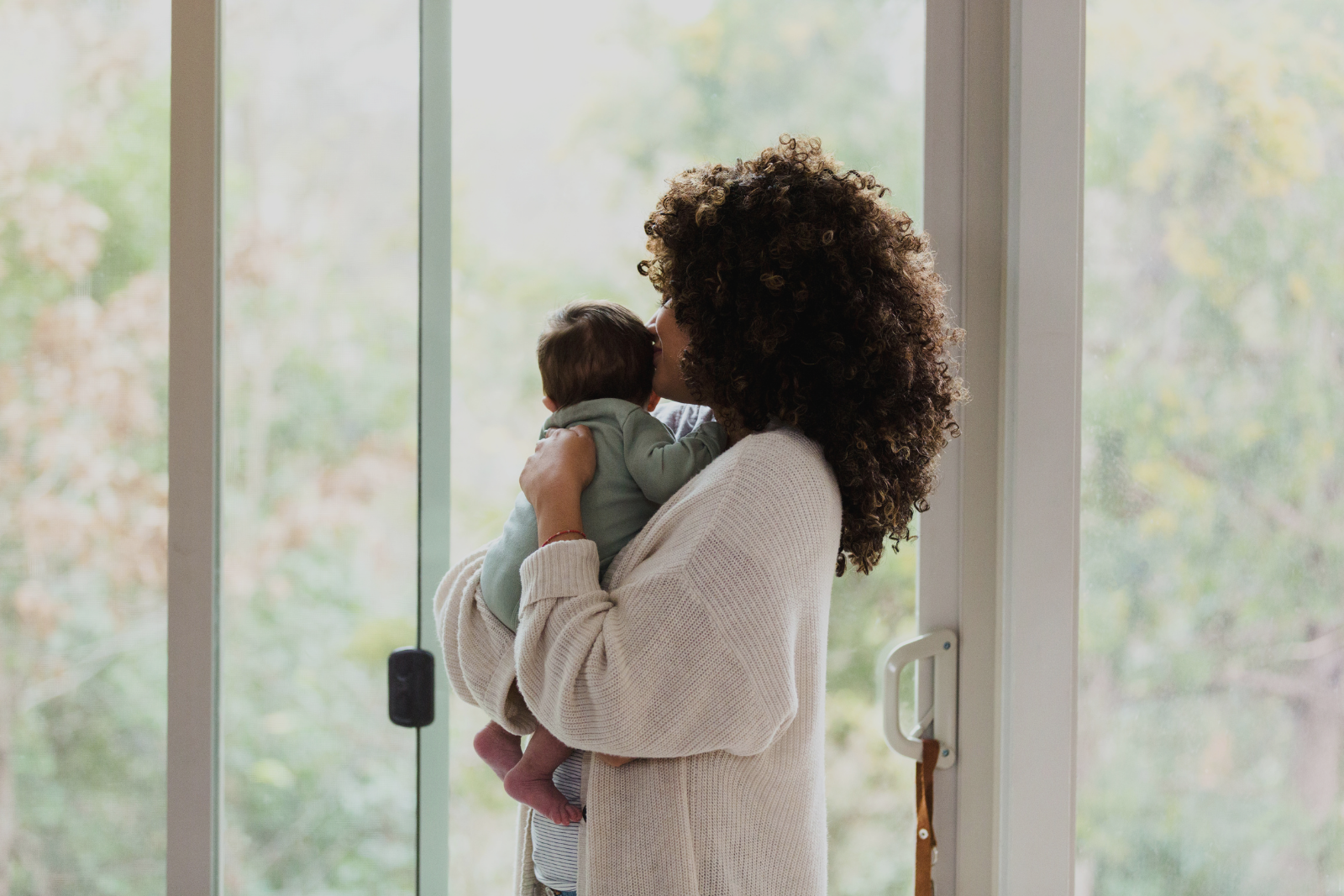 A woman with curly hair holding a baby near a large window in her Bend Oregon home, after her in-home lactation consultation and IBCLC newborn support Bend OR