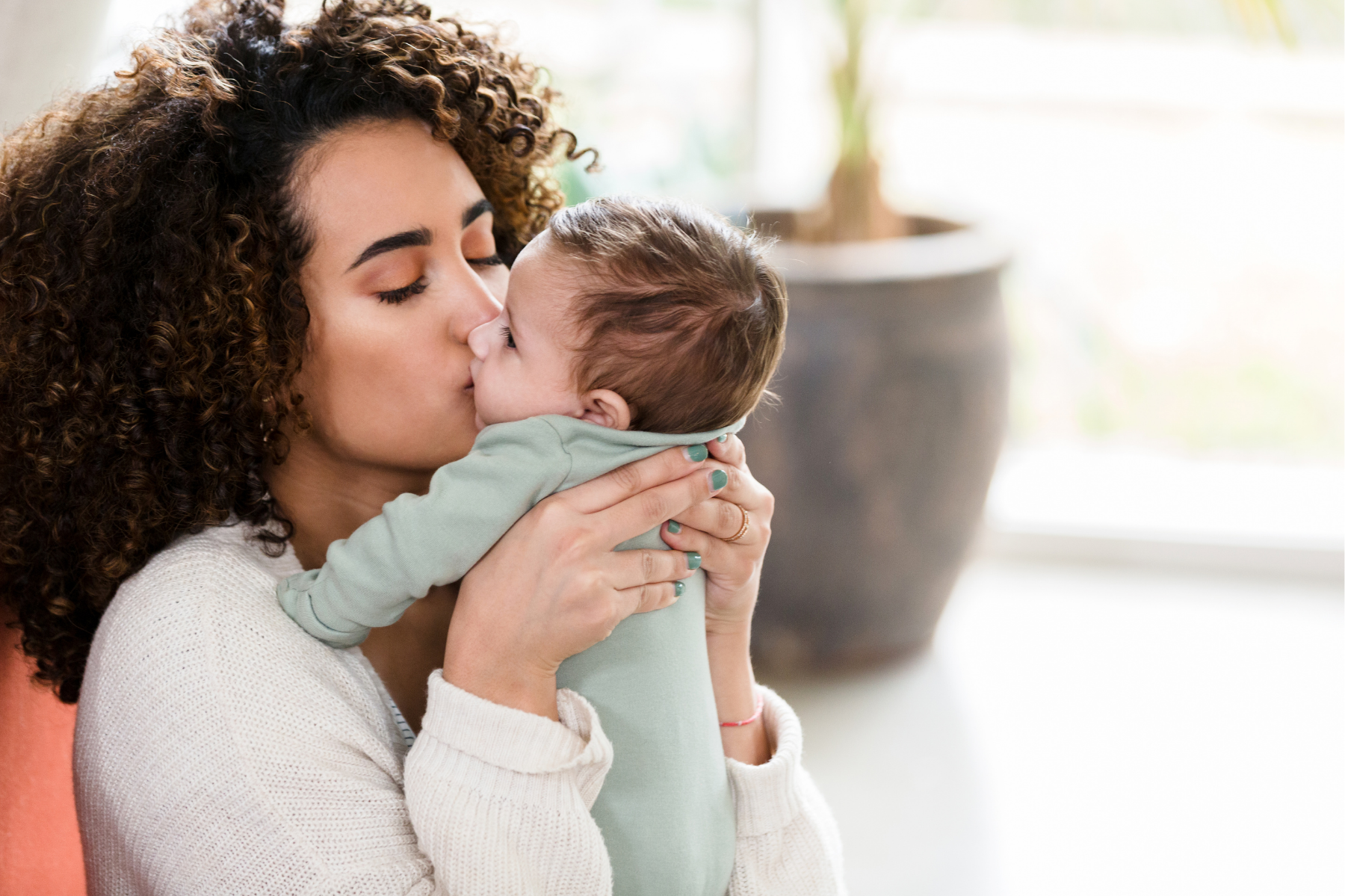 A woman with curly hair holds and kisses a young boy on the cheek, after a calm infant massage practice promoting connection and soothing care for her baby