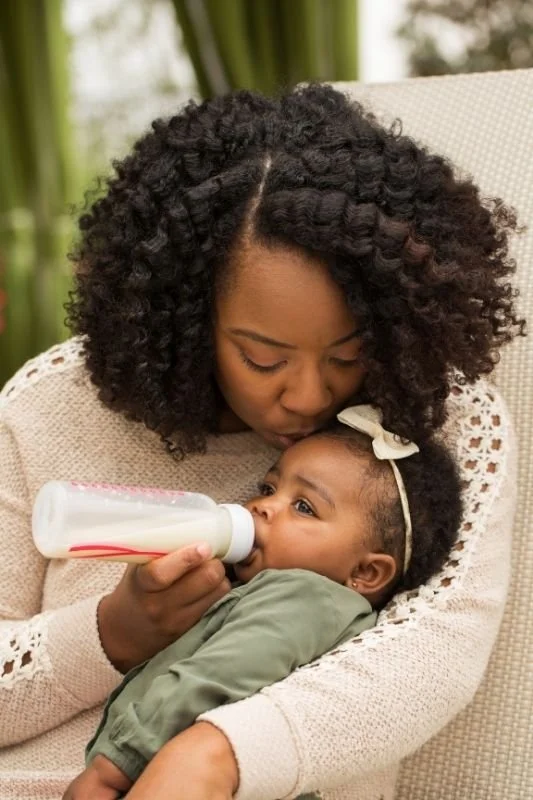 Woman feeding a baby with a bottle while sitting on a beige chair, in her home in Bend, Oregon.