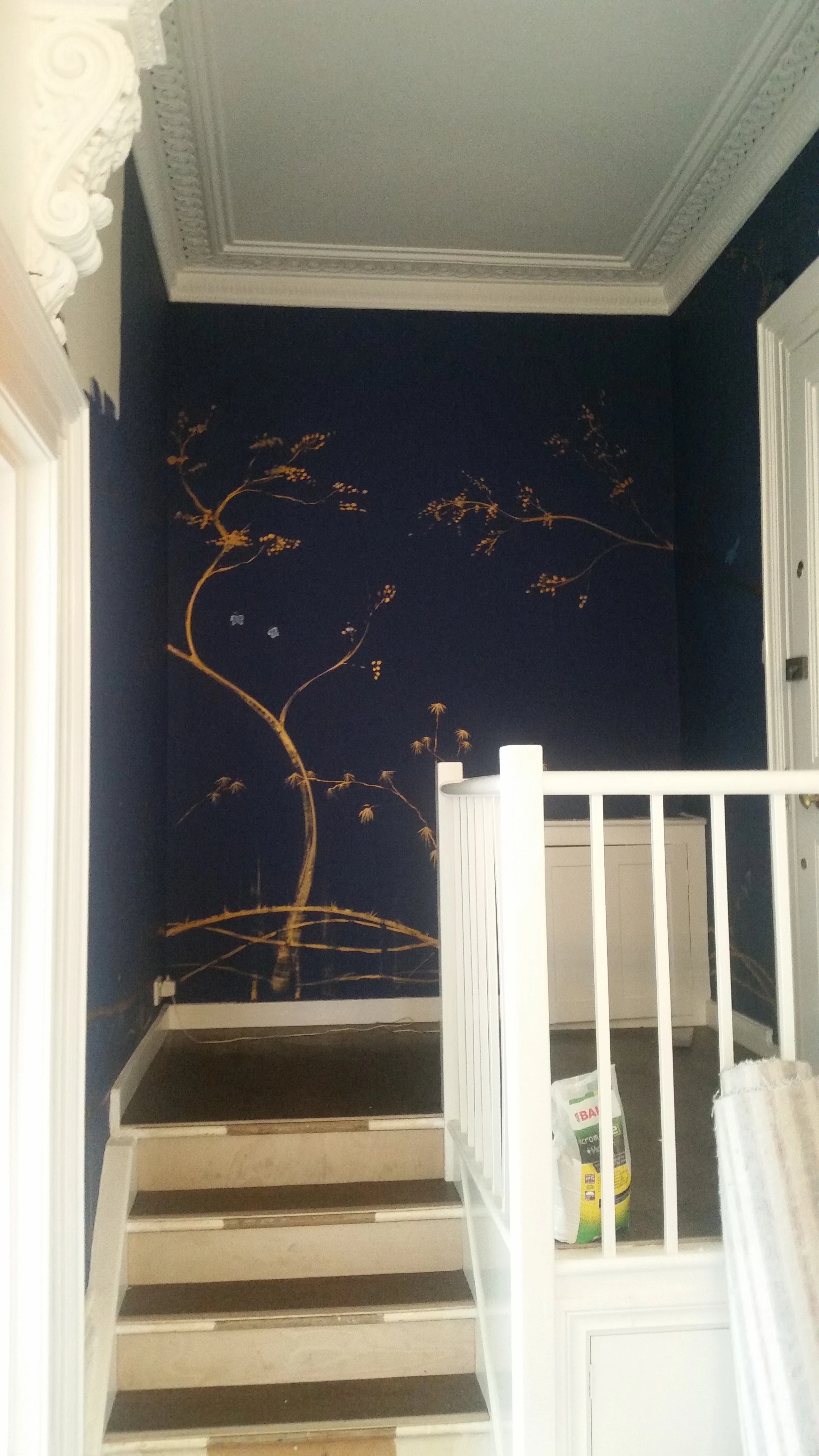 Interior view of a staircase with a dark blue mural featuring a gold leaf design, white crown molding ceiling, and white railings.