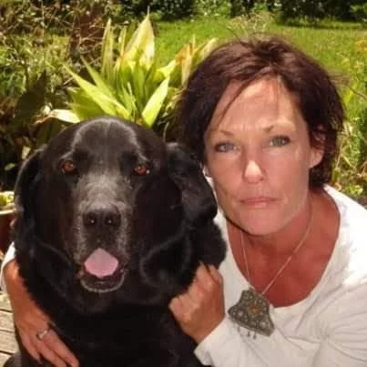 Helena with a large black dog outdoors with green plants in the background.