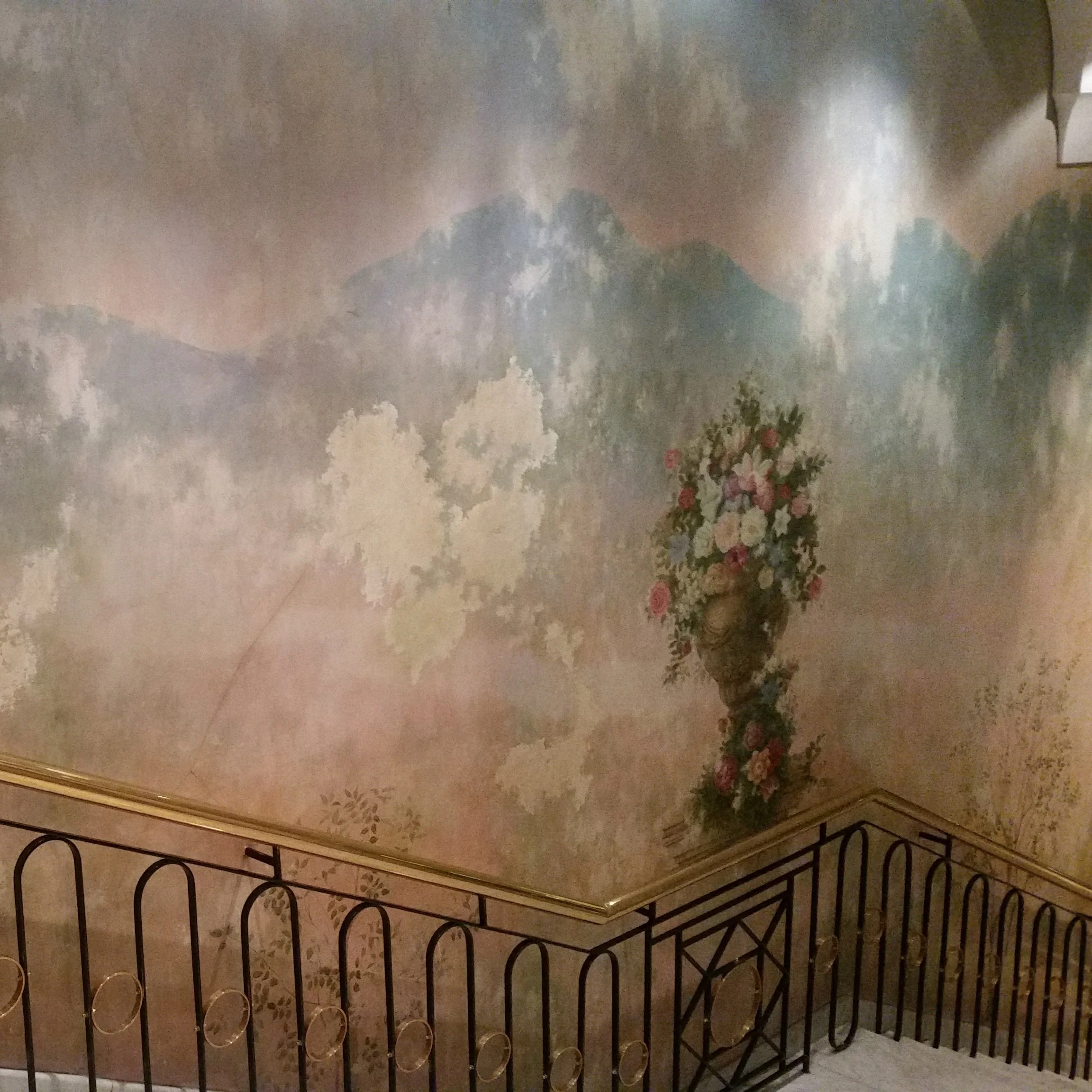 Wall mural with mountains, clouds, and a floral vase on a pedestal in a room with a decorative black metal railing.