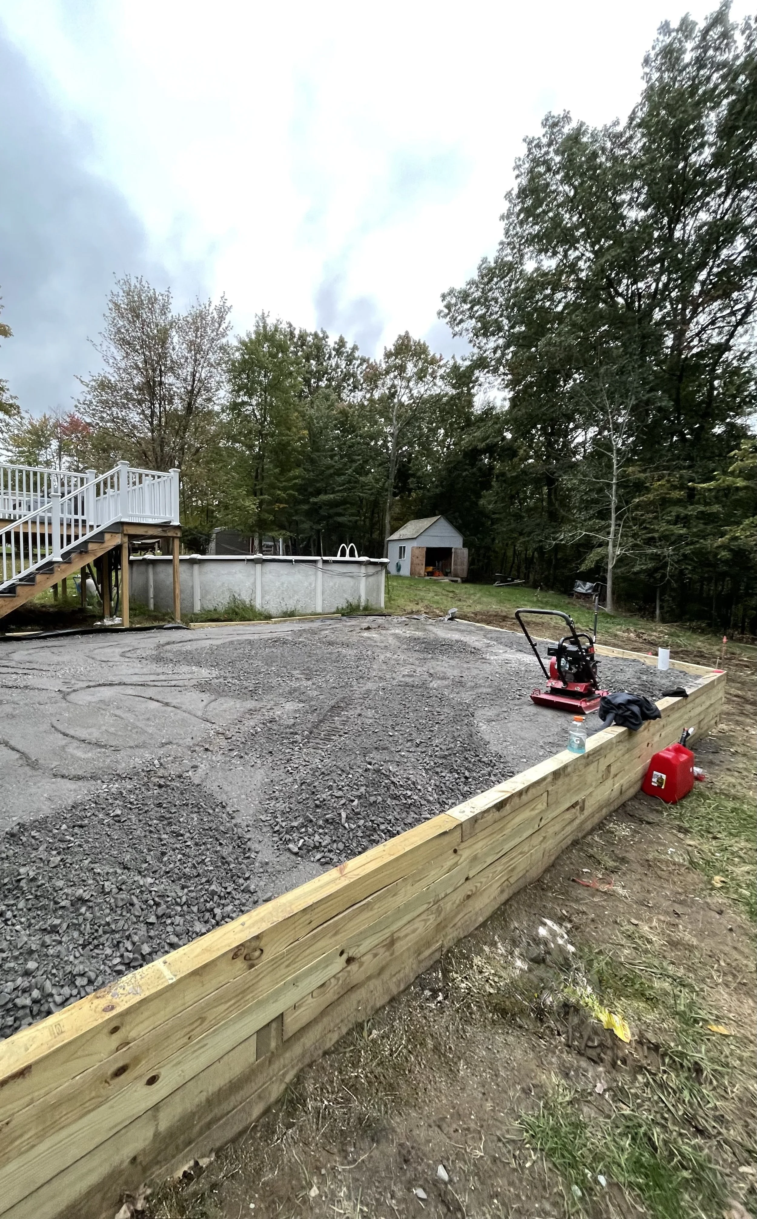 Gravel-covered area with wooden retaining wall, surrounded by trees, next to an above-ground pool and a shed, with construction equipment and supplies visible.