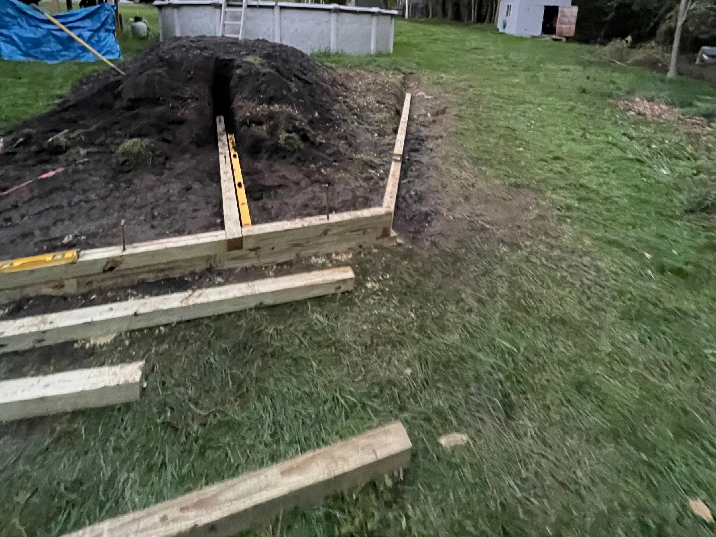 Wooden frames and dirt pile with trench on grassy lawn, near above-ground pool and shed.