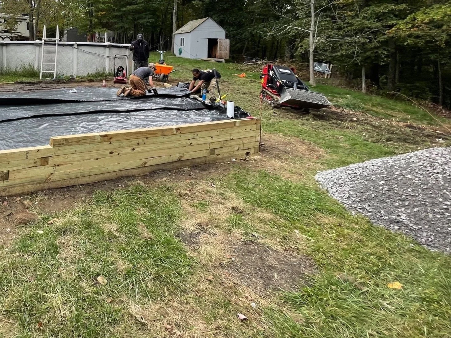 People working on a backyard landscaping project with a mini excavator and a wooden frame structure.