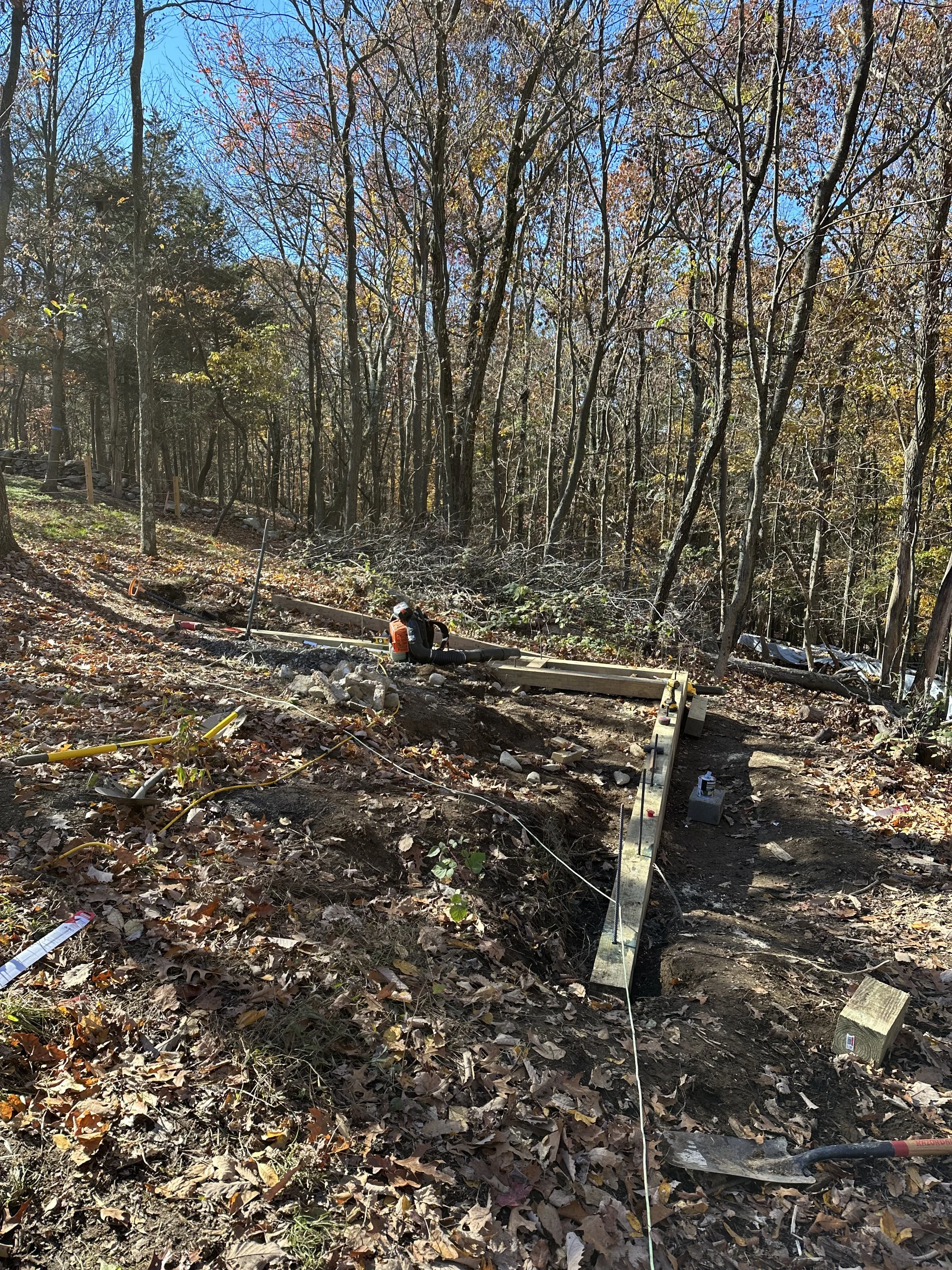 Outdoor construction site in a wooded area with scattered leaves, showcasing wooden beams and construction tools on the ground.