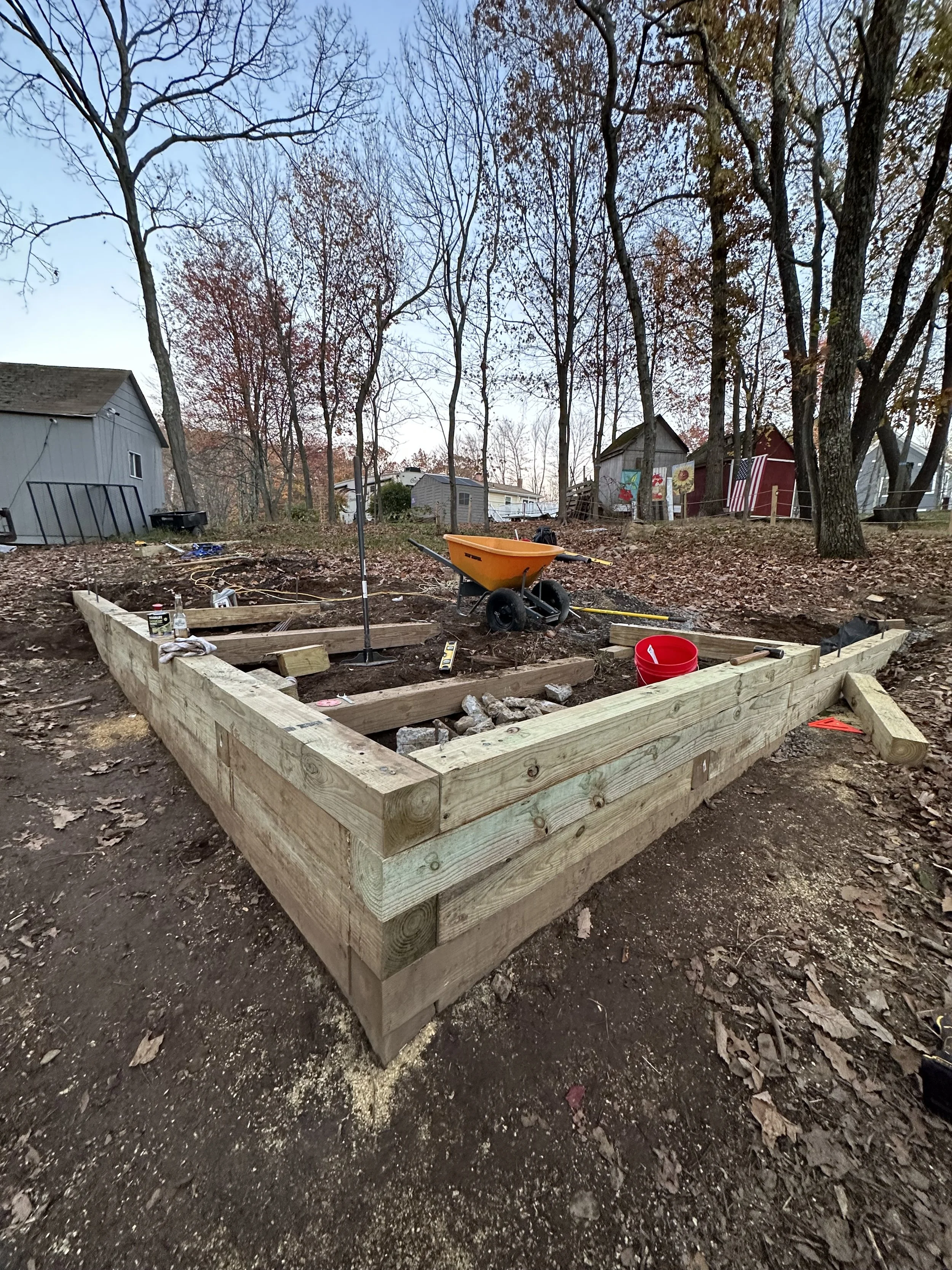Wooden foundation under construction in a wooded area, surrounded by trees and fallen leaves, with a wheelbarrow and a red bucket nearby.