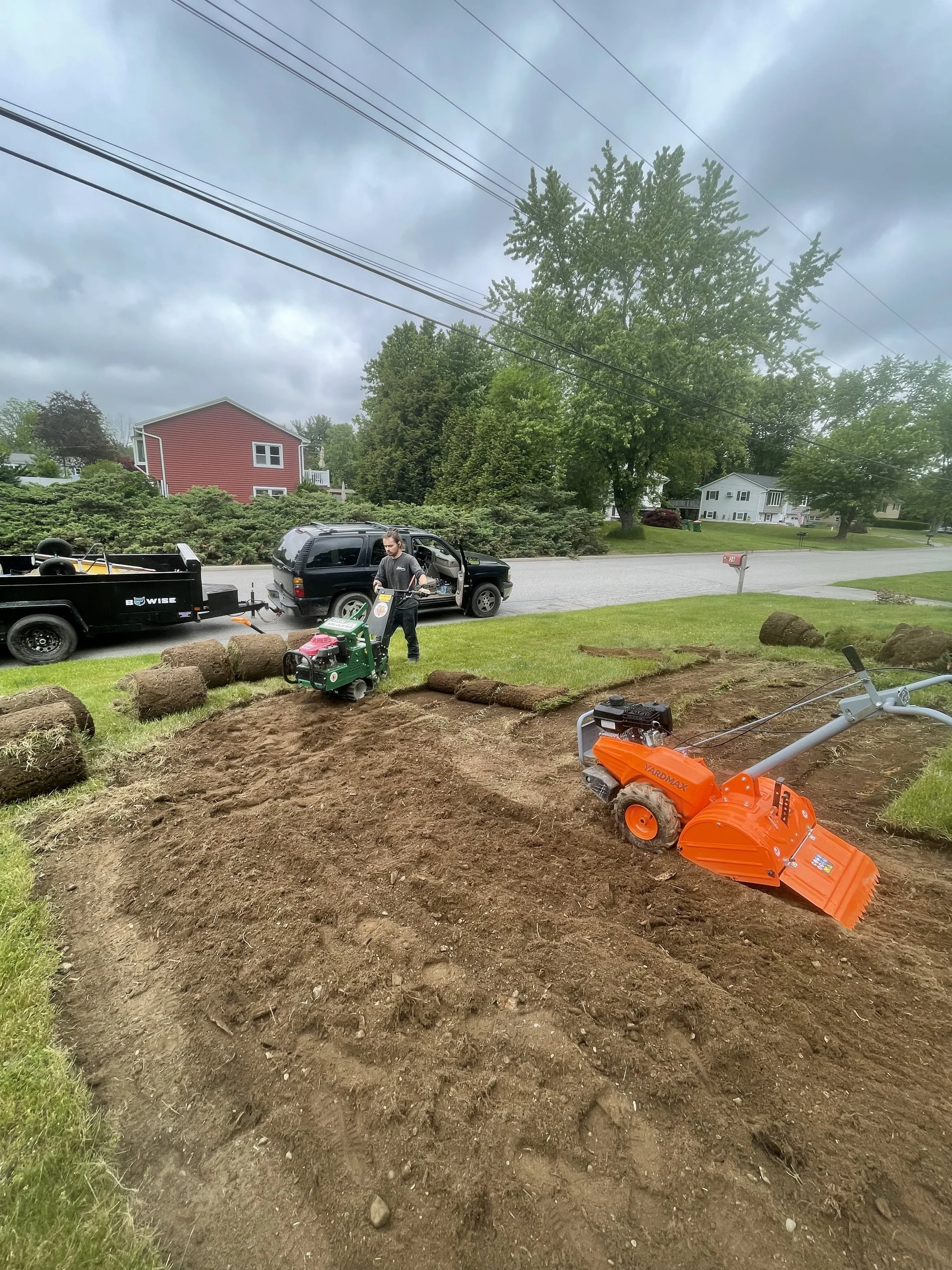 Person working on lawn installation with gardening equipment, sod rolls, and tiller in suburban yard.