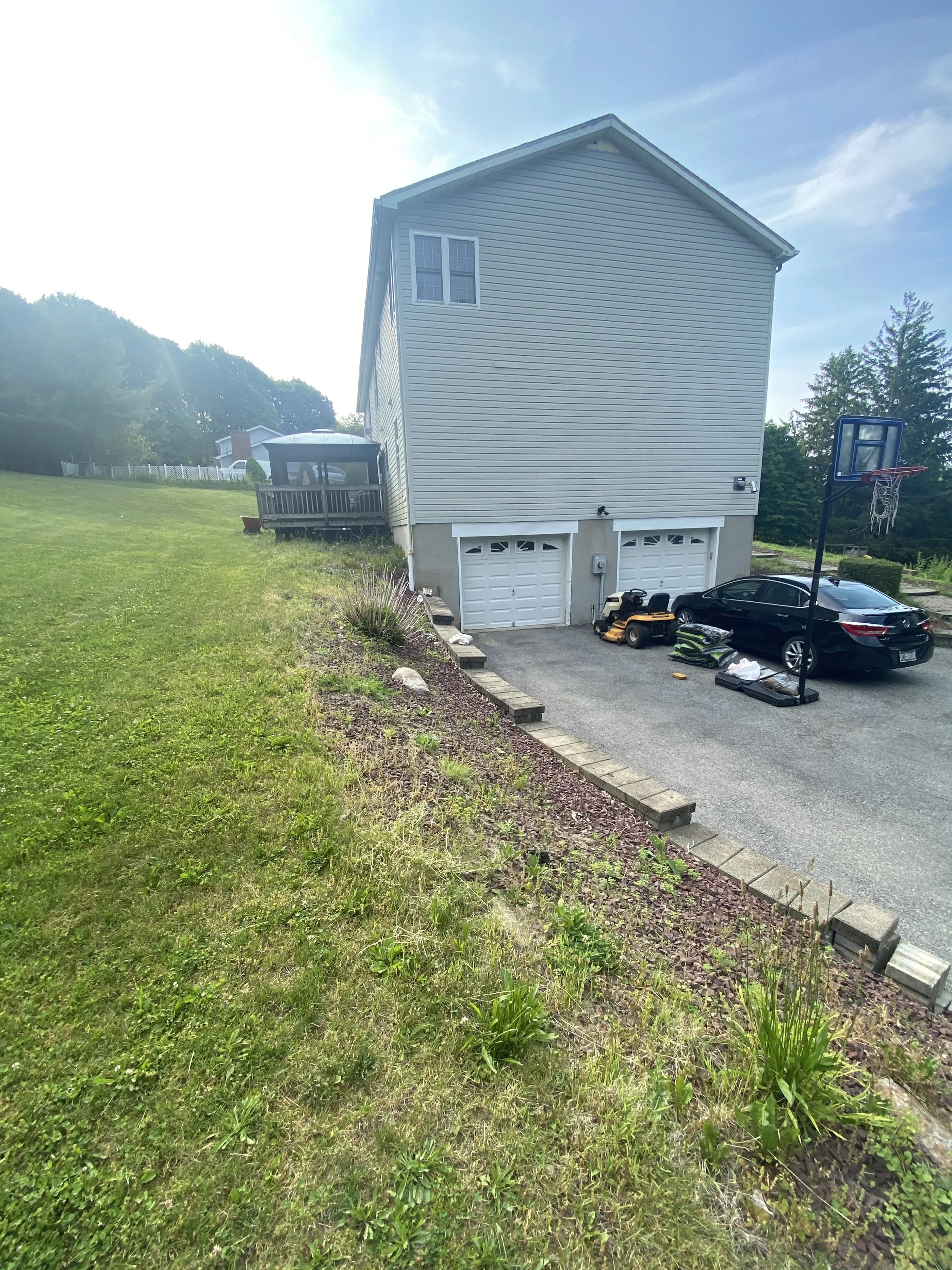 Side view of a house with two garage doors, a driveway with a parked car, and a basketball hoop; adjacent grassy lawn and trees in the background.