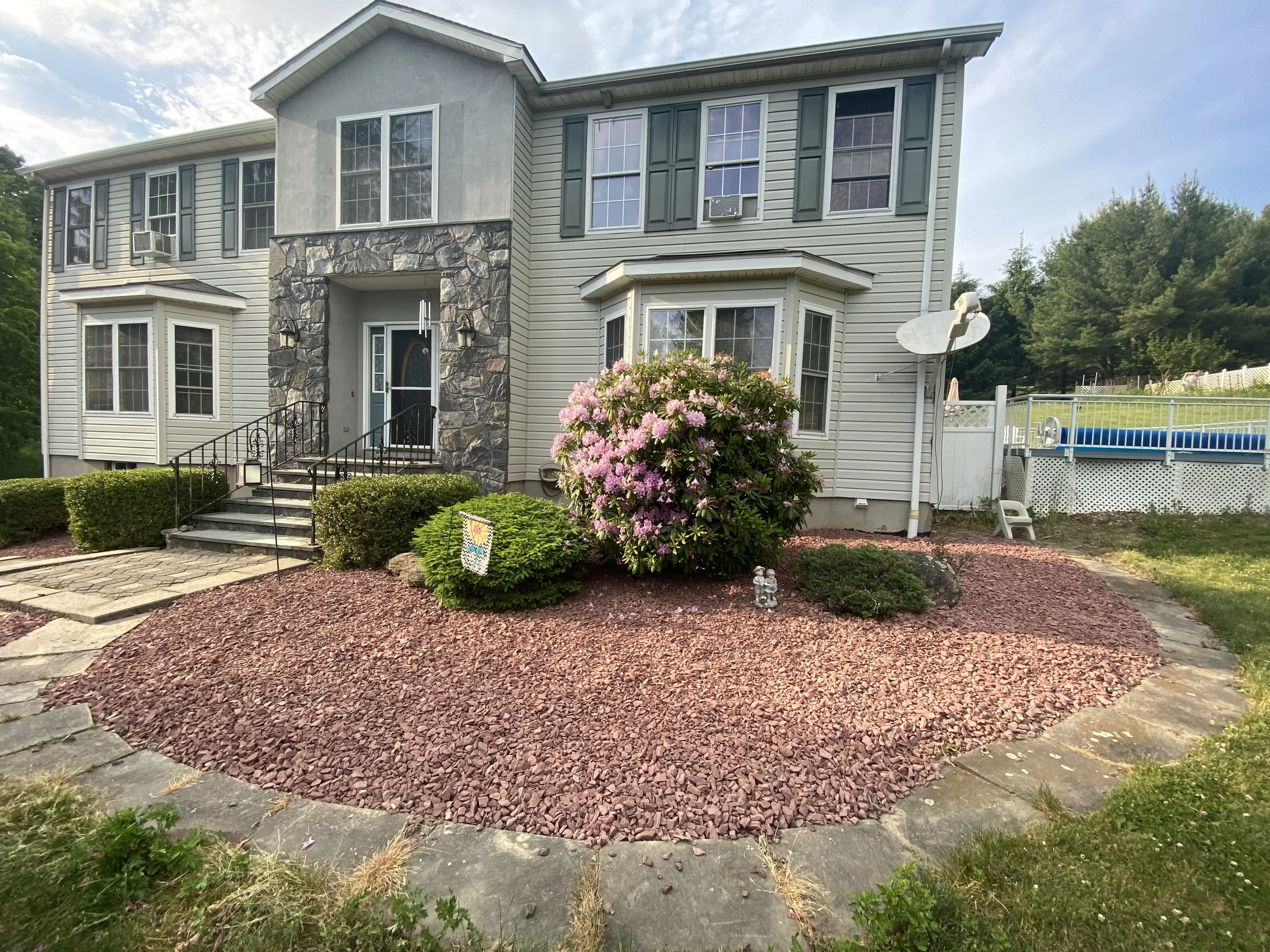 Two-story house with stone and siding exterior, surrounded by landscaped garden with pink flowering bush and red mulch. A satellite dish is attached to the side, and a rectangular swimming pool is visible in the backyard.