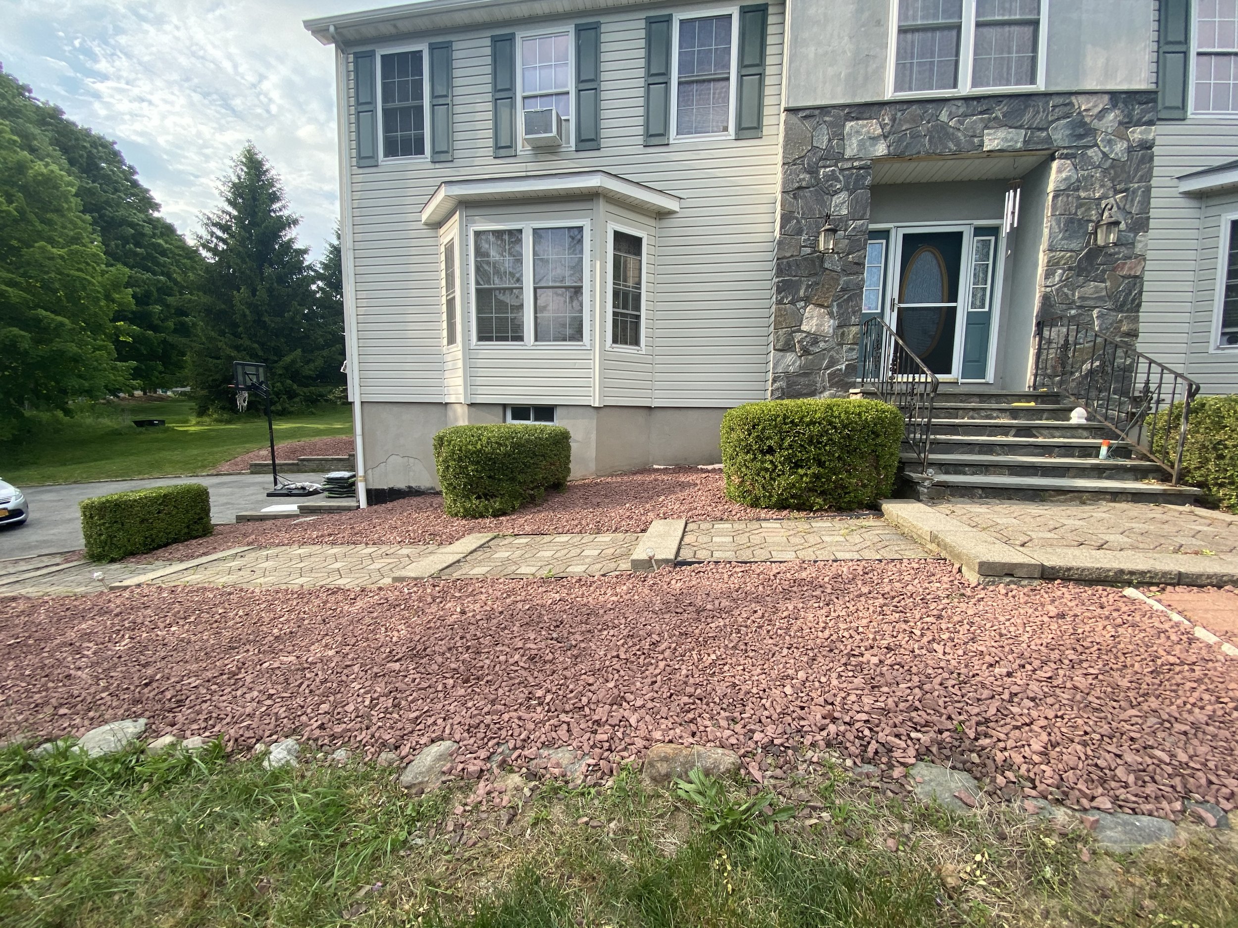 Front entrance of a two-story house with stone and siding exterior, steps leading up to a front door, landscaped with red gravel and green shrubs, driveway with a basketball hoop on the side.