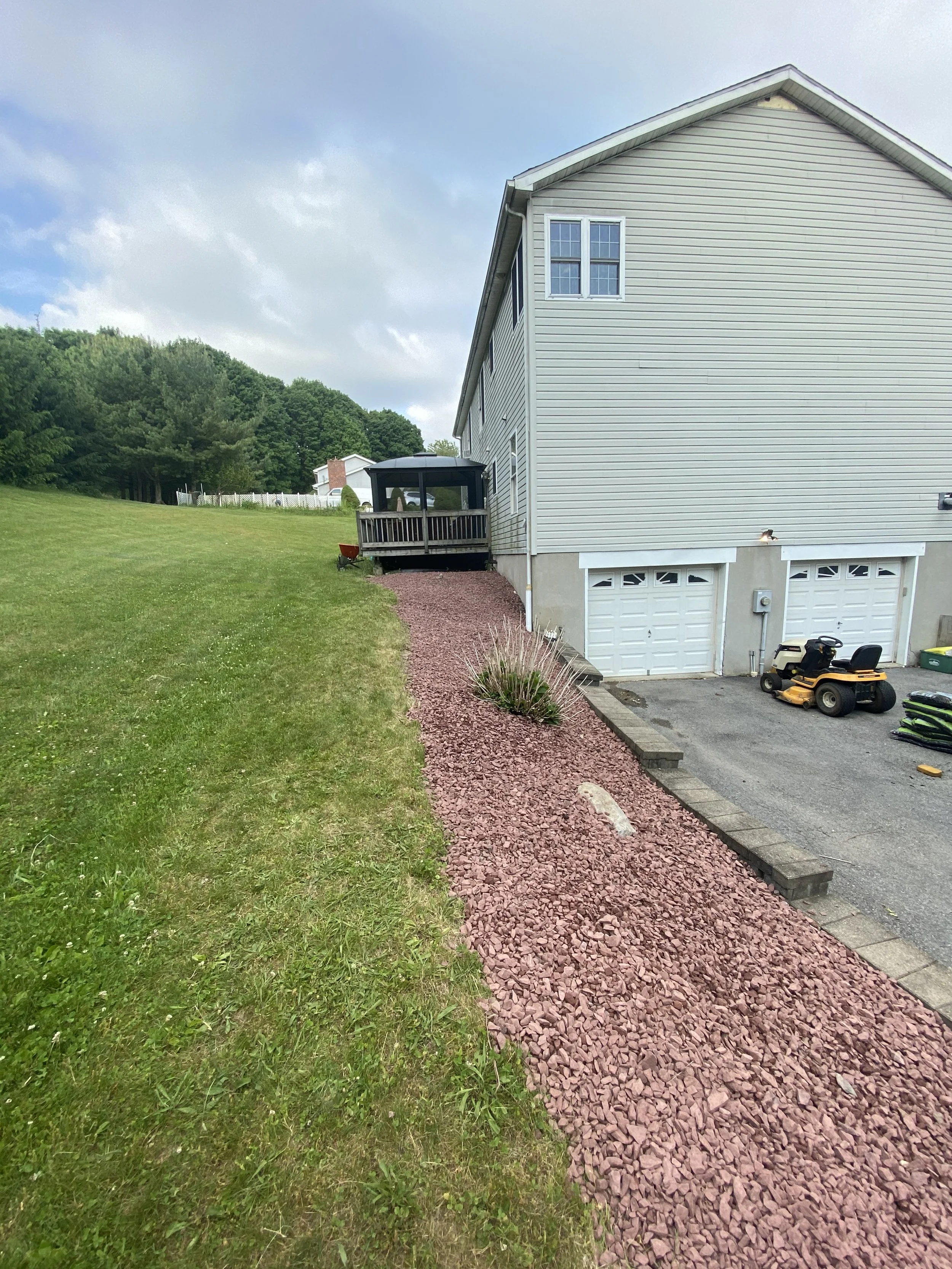 Side view of a house with a gravel path and a deck, surrounded by a grassy area and trees in the background. Two garage doors and a lawn mower are visible.