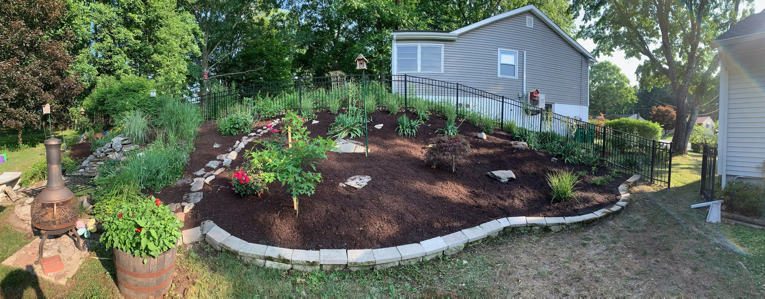 A landscaped garden with various plants, shrubs, and flowers, surrounded by a curved stone border. The garden is on a slope next to a house with light siding. There is a black metal fence along the slope. A barrel planter with red flowers and a chiminea are in the foreground. Tall trees and a neighboring house are visible in the background.