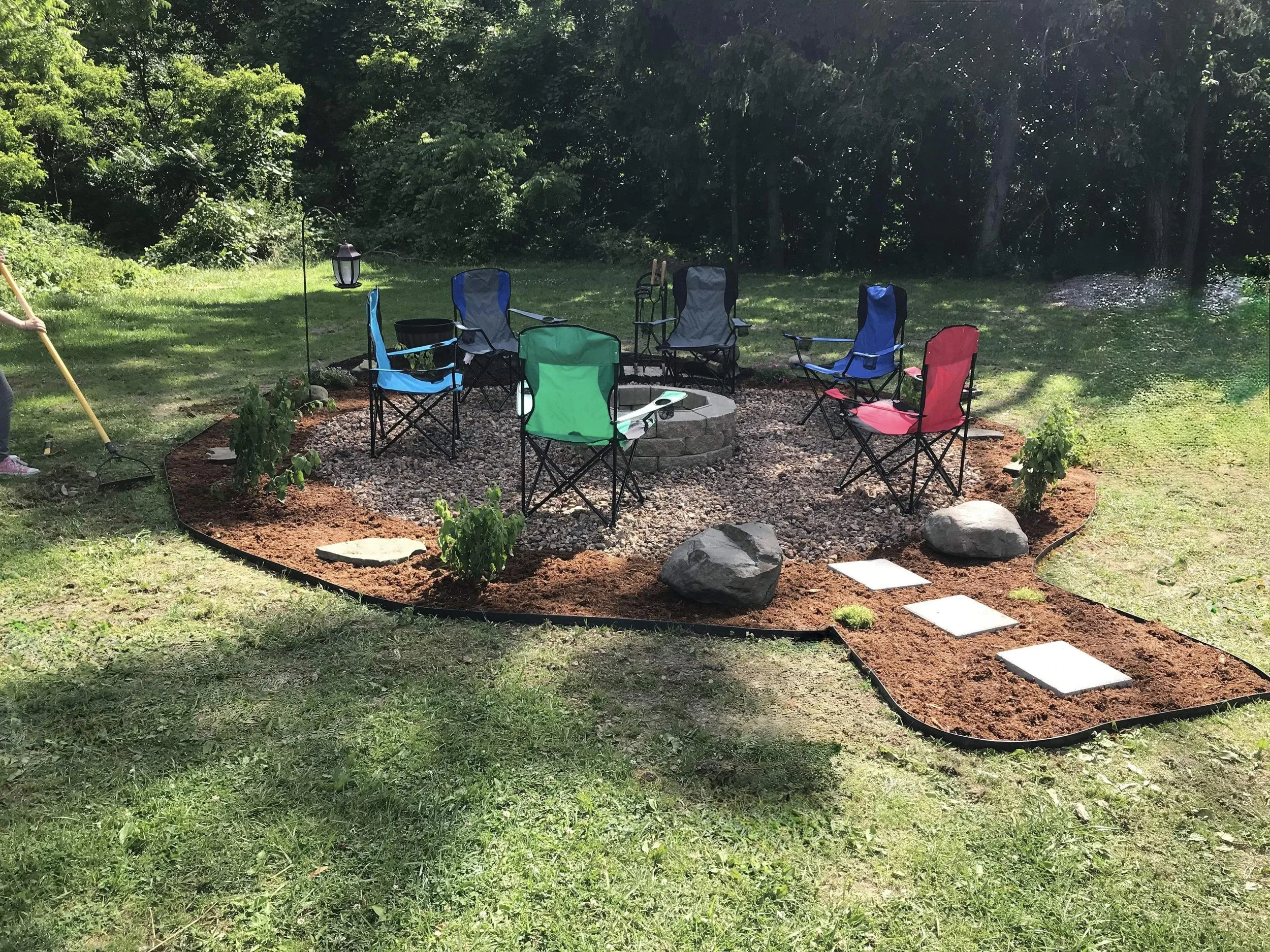 Outdoor fire pit area with gravel, surrounded by colorful folding chairs, mulch, stone pathway, and greenery.